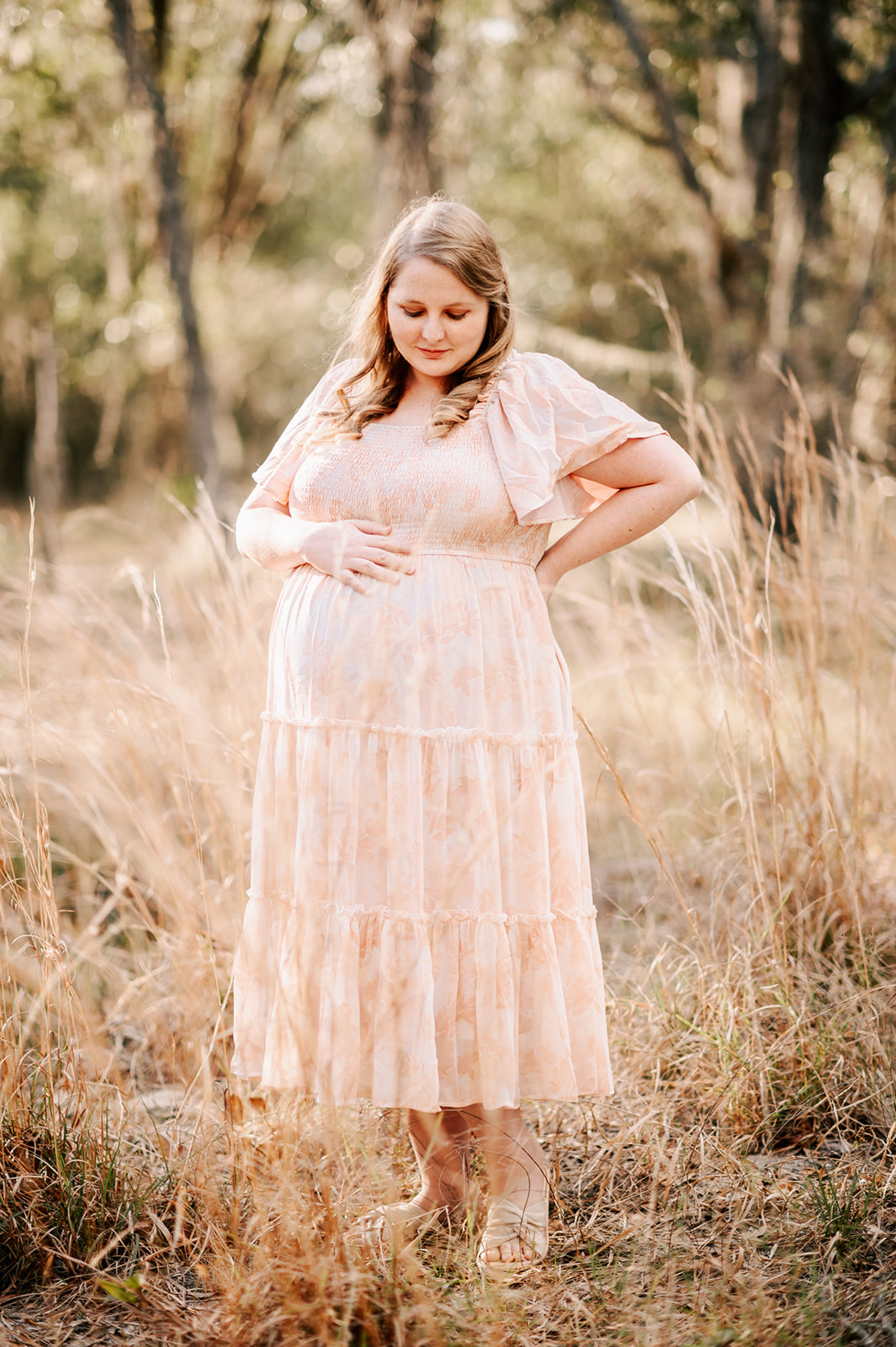 A pregnant woman stands in a field of golden grass while looking down at her bump in a pink floral dress