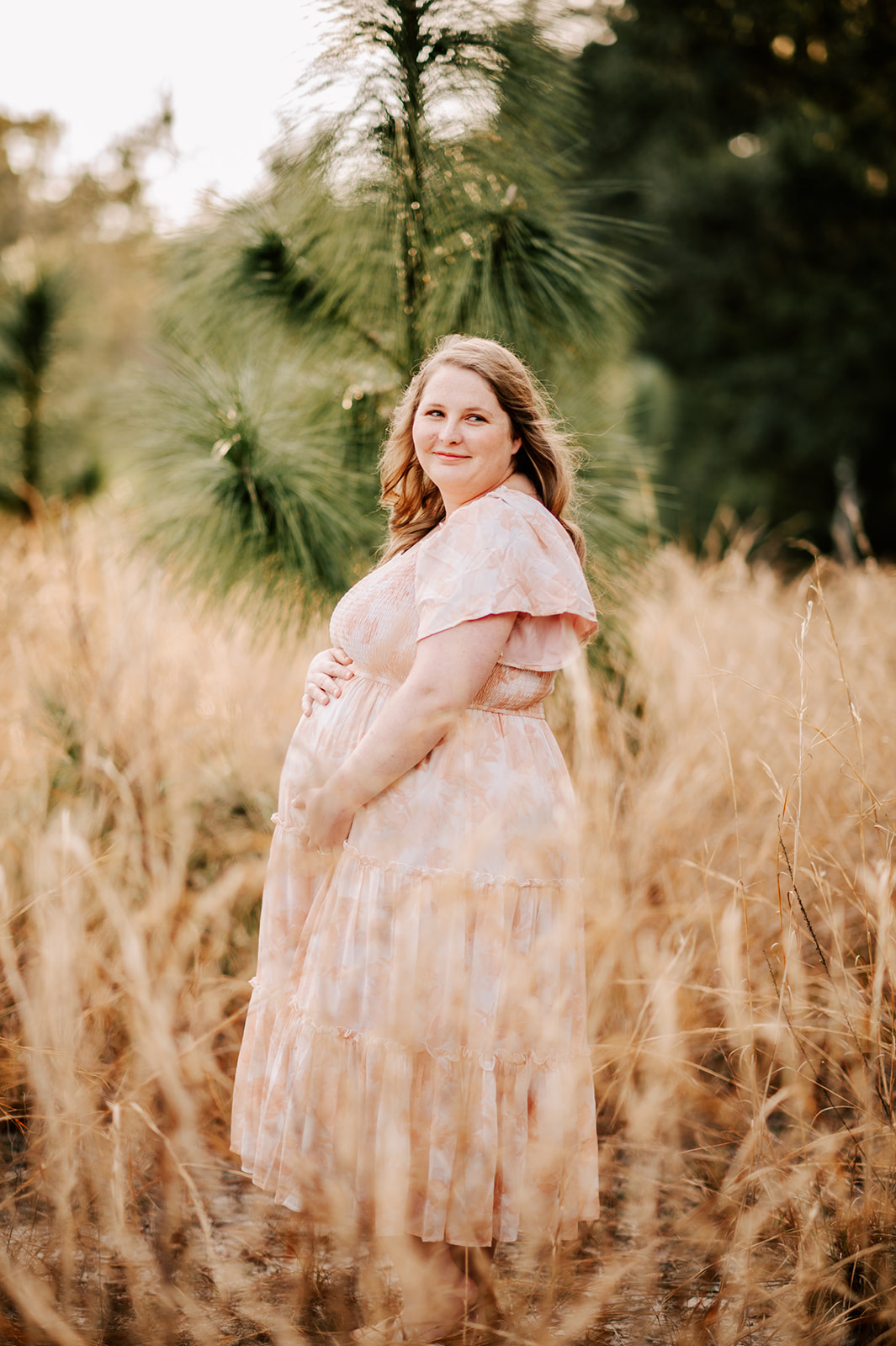 A mom-to-be stands in a field of golden grass while holding her bump with both hands in a pink maternity dress Durham Womens Clinic
