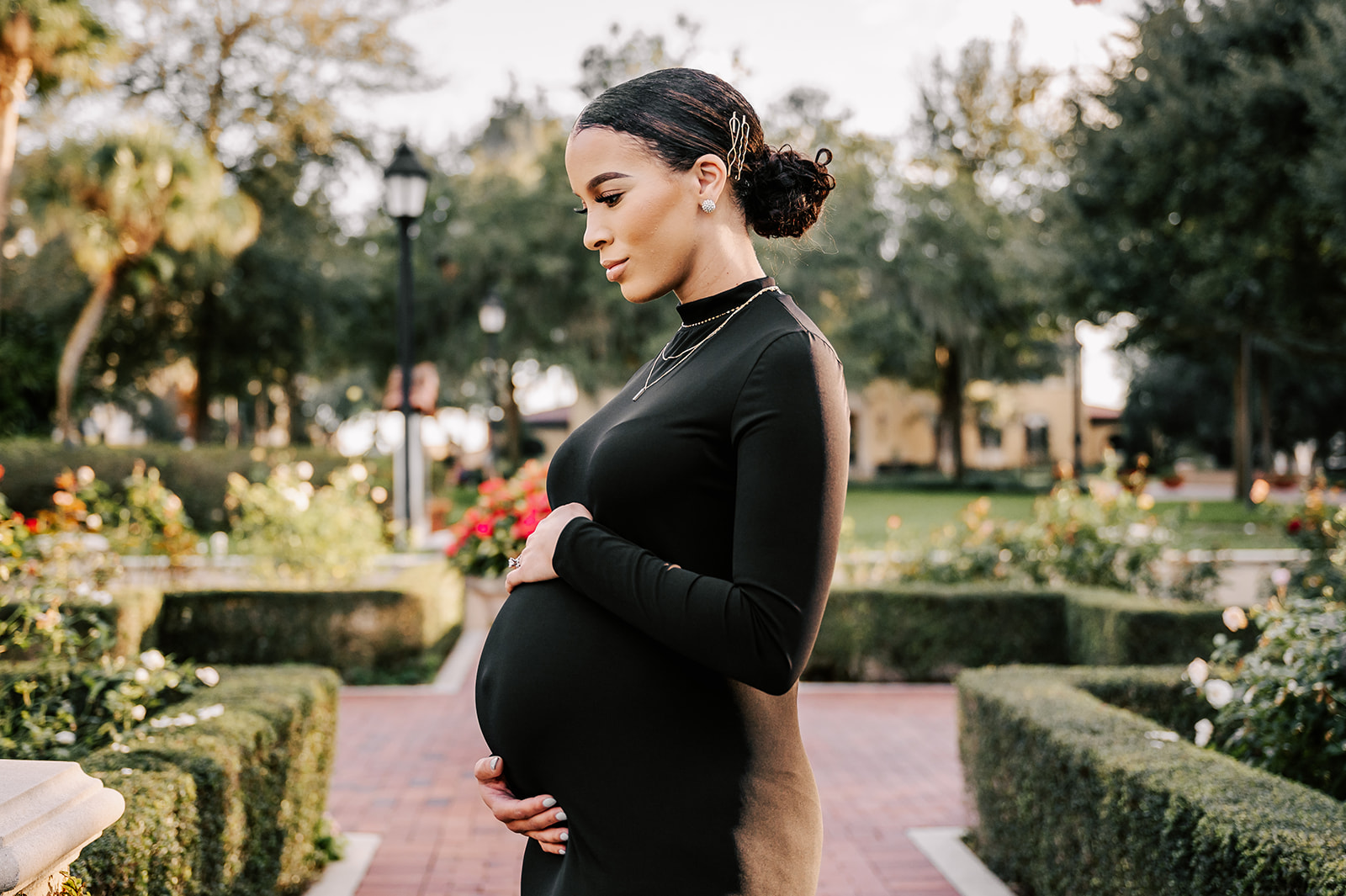 A pregnant woman stands on a brick path in a rose garden while holding the top and bottom of her bump