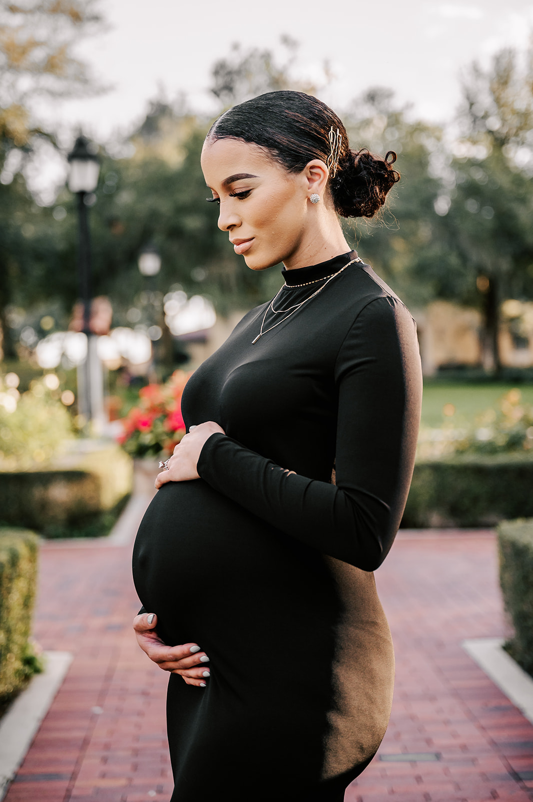 A pregnant woman stands on a brick path in a rose garden while holding the top and bottom of her bump Precious Arrrows
