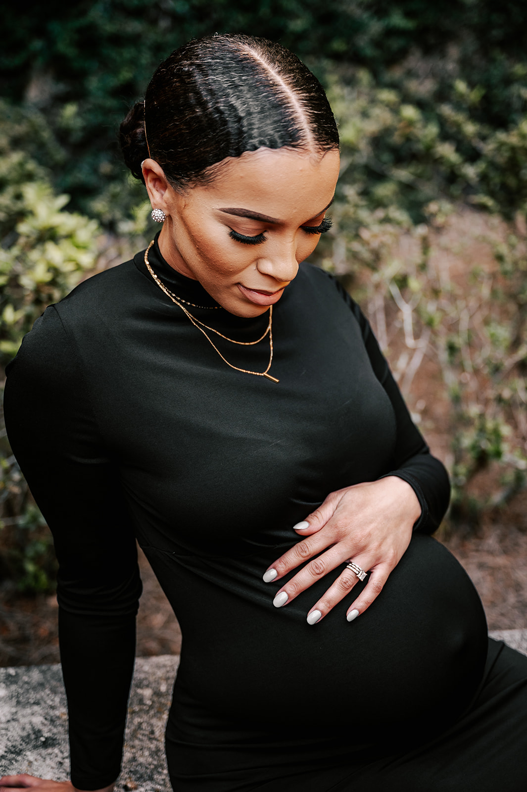 A mom to be in a black maternity gown sits on a rock in the woods while looking down at her bump Precious Arrrows