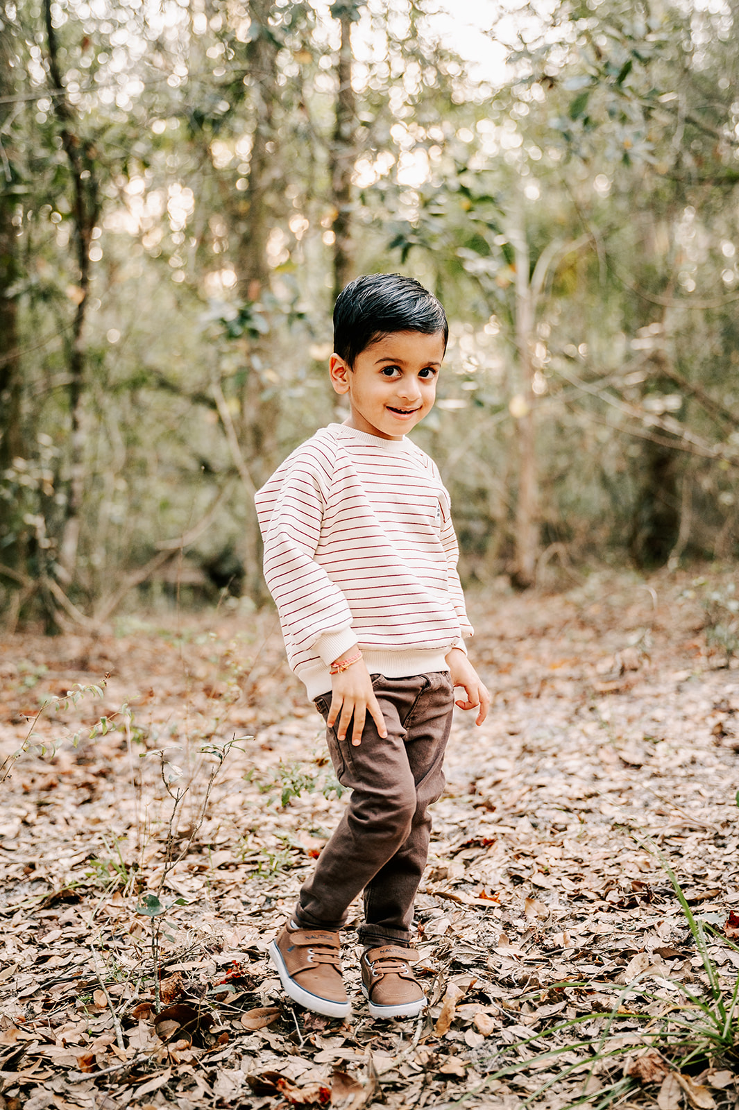 A young boy in brown pants and a stripe shirt stands in a forest with thumbs in his pockets