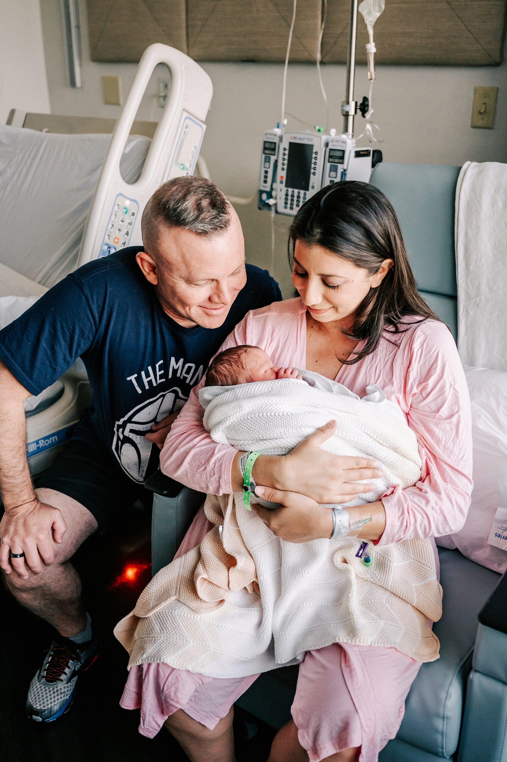 New parents sit in a hospital room chair looking down at their newborn baby sleeping in a bundle of blanket in mom's arms advance community health apex