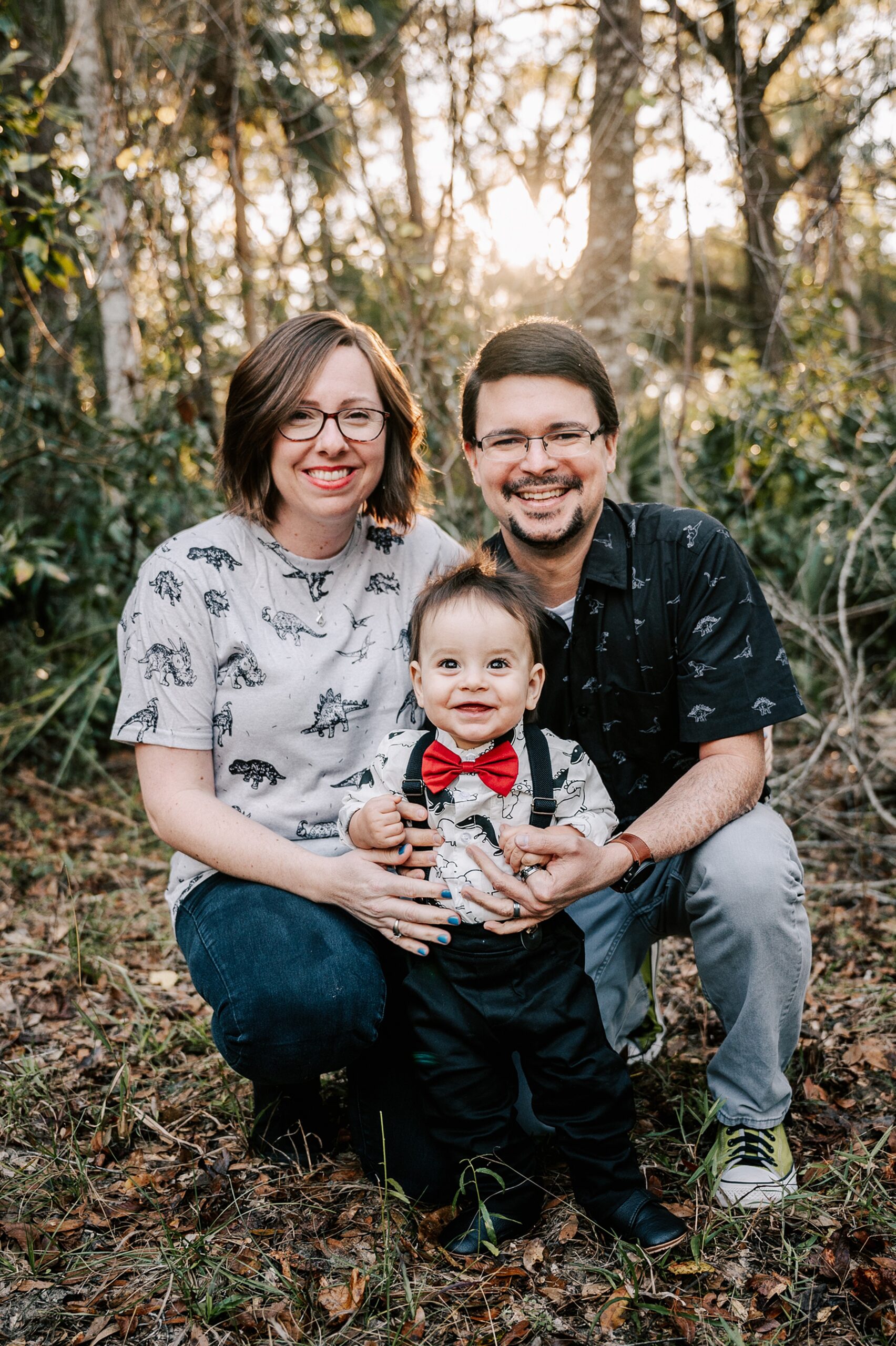 Happy parents kneel in a forest with their toddler son standing between them