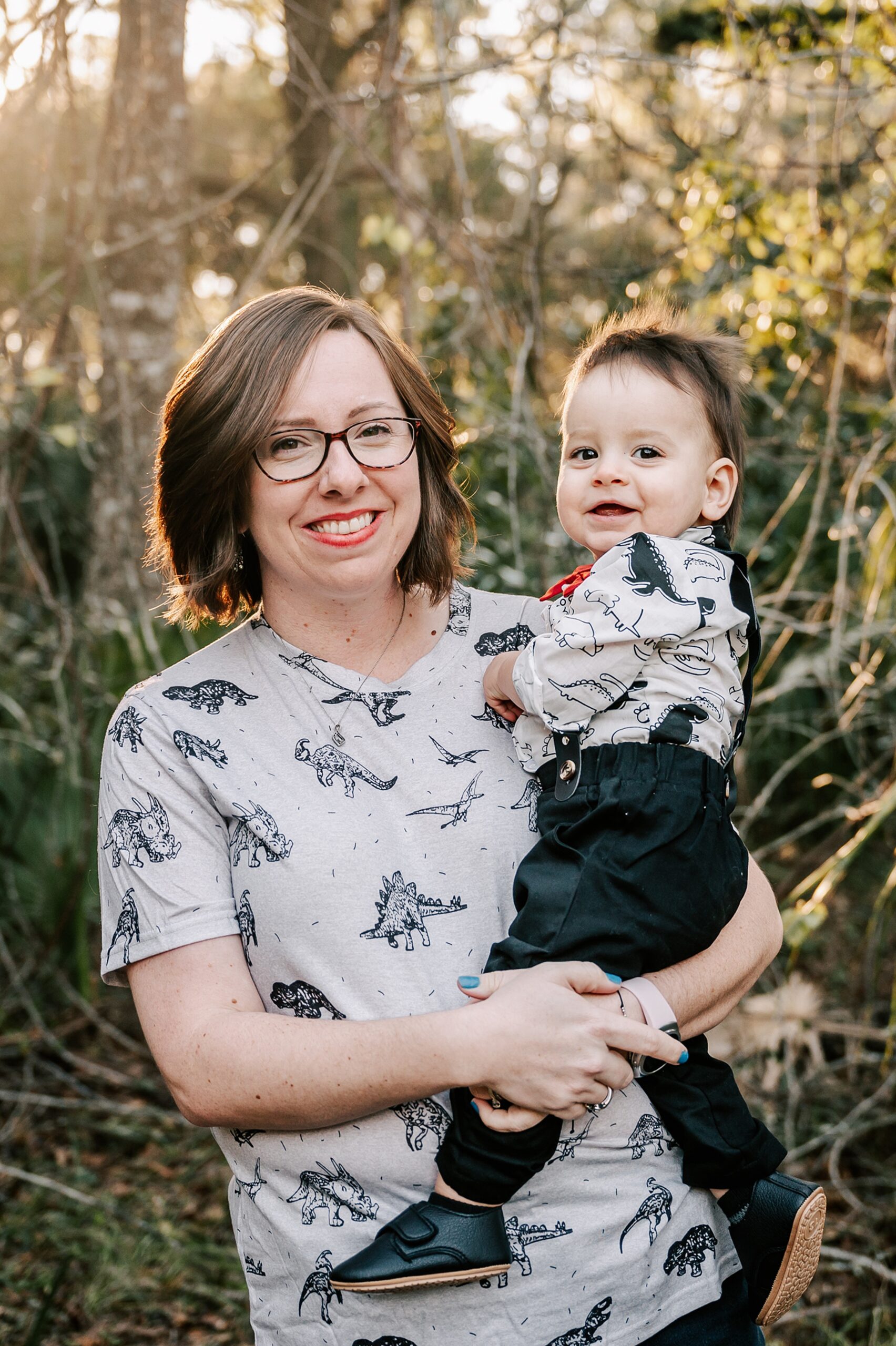 A mother in a dinosaur shirt stands in a forest holding her toddler son in matching shirt and red bowtie on her hip ardmore baptist preschool