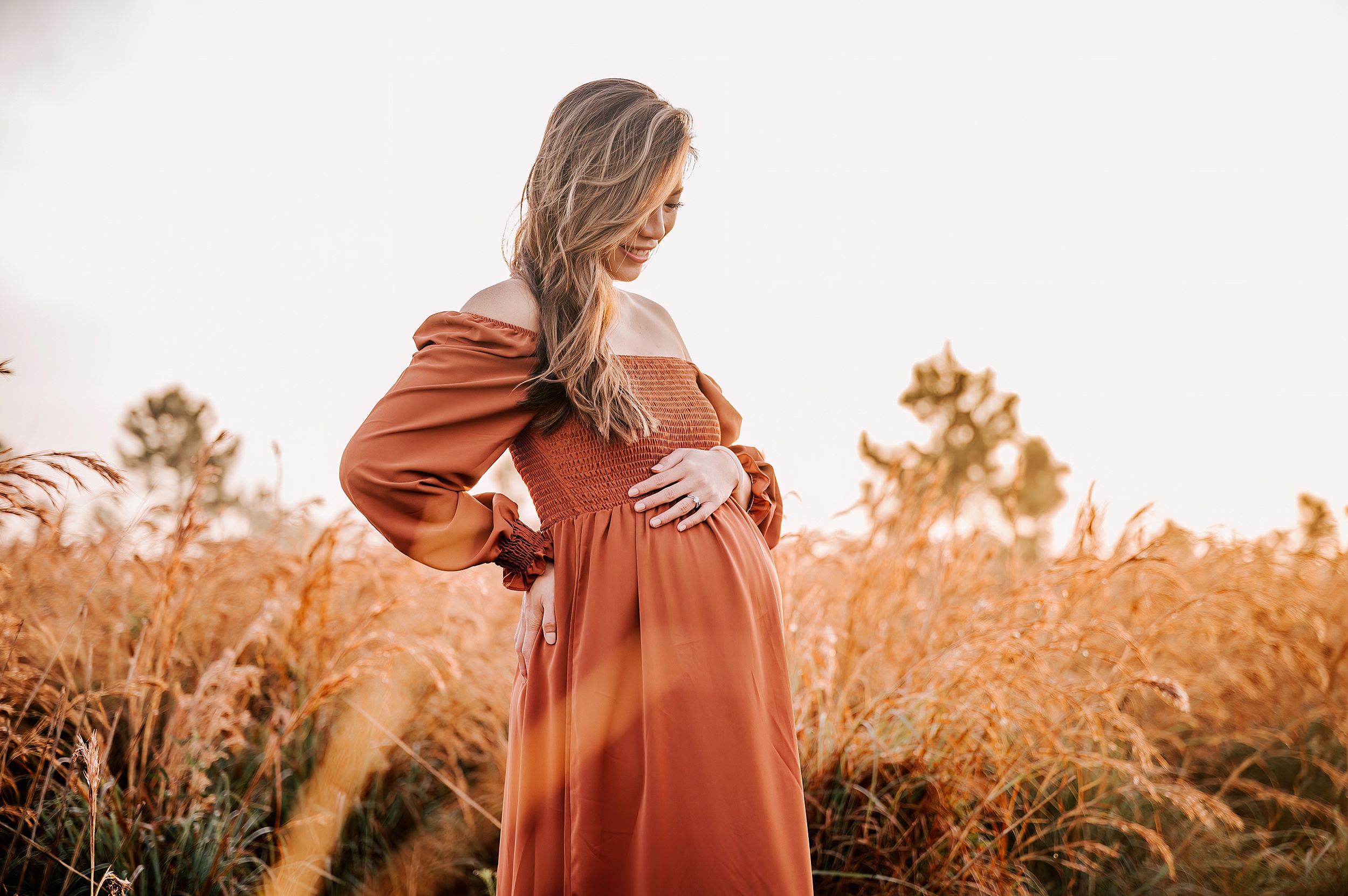 A mother to be smiles down at her bump while standing iin a field of golden grass at sunrise