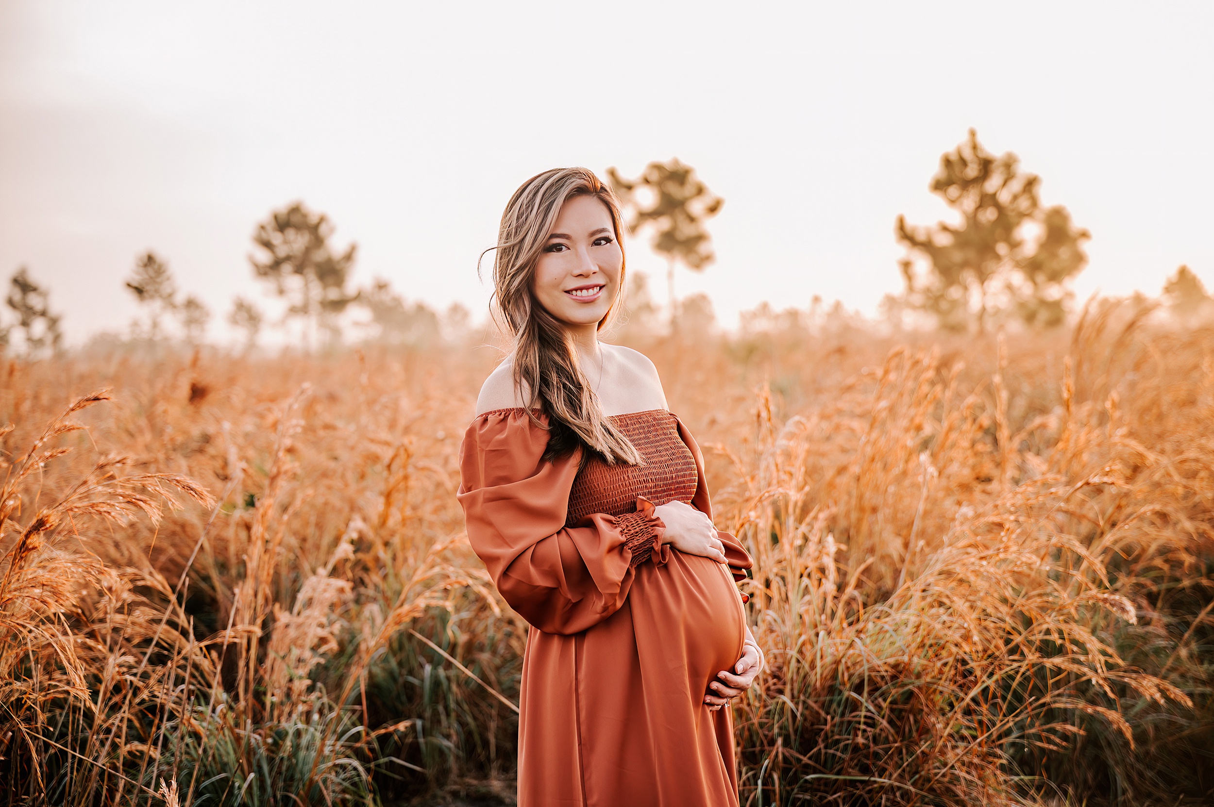 A mother to be in a red maternity gown stands in a field of tall golden grass at sunrise while holding her bump Bella Labor