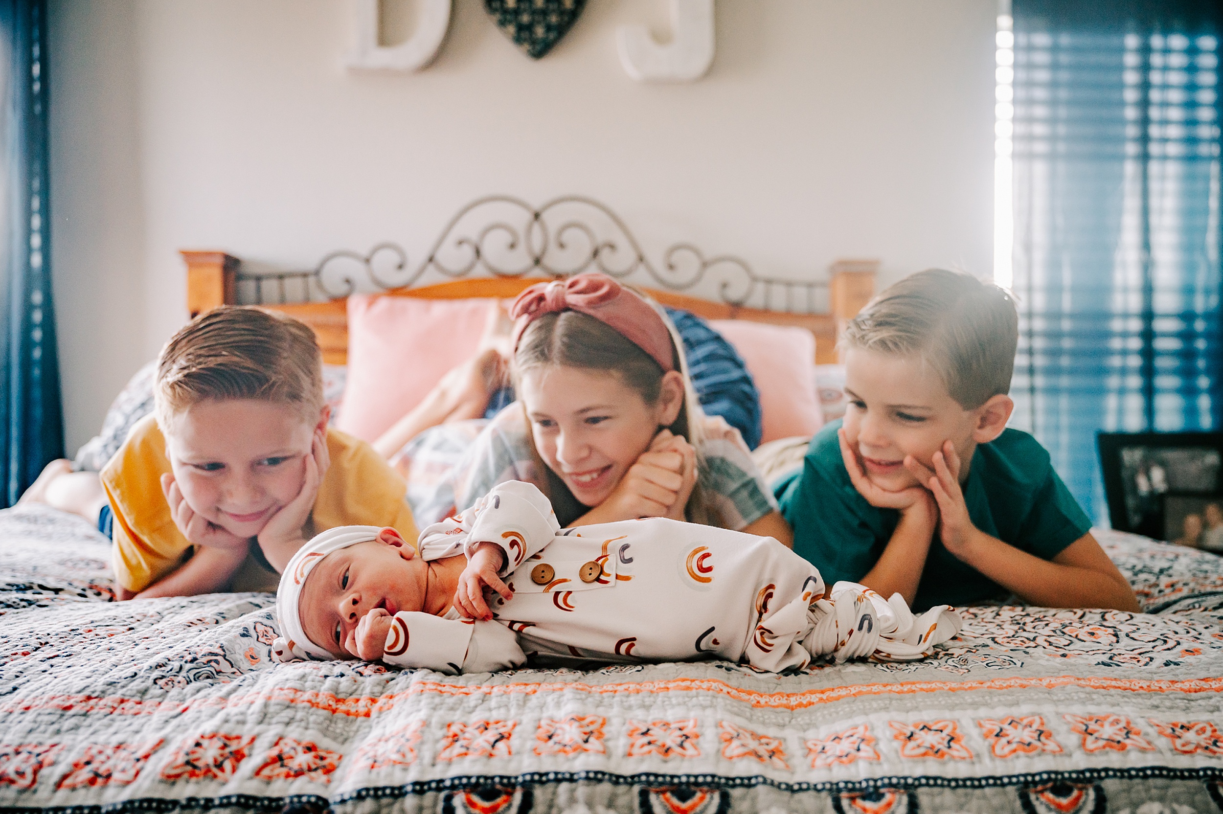 Three siblings lay on a bed resting their heads in their hands while looking at their newborn baby sibling laying across the end of the bed brookhaven country day school