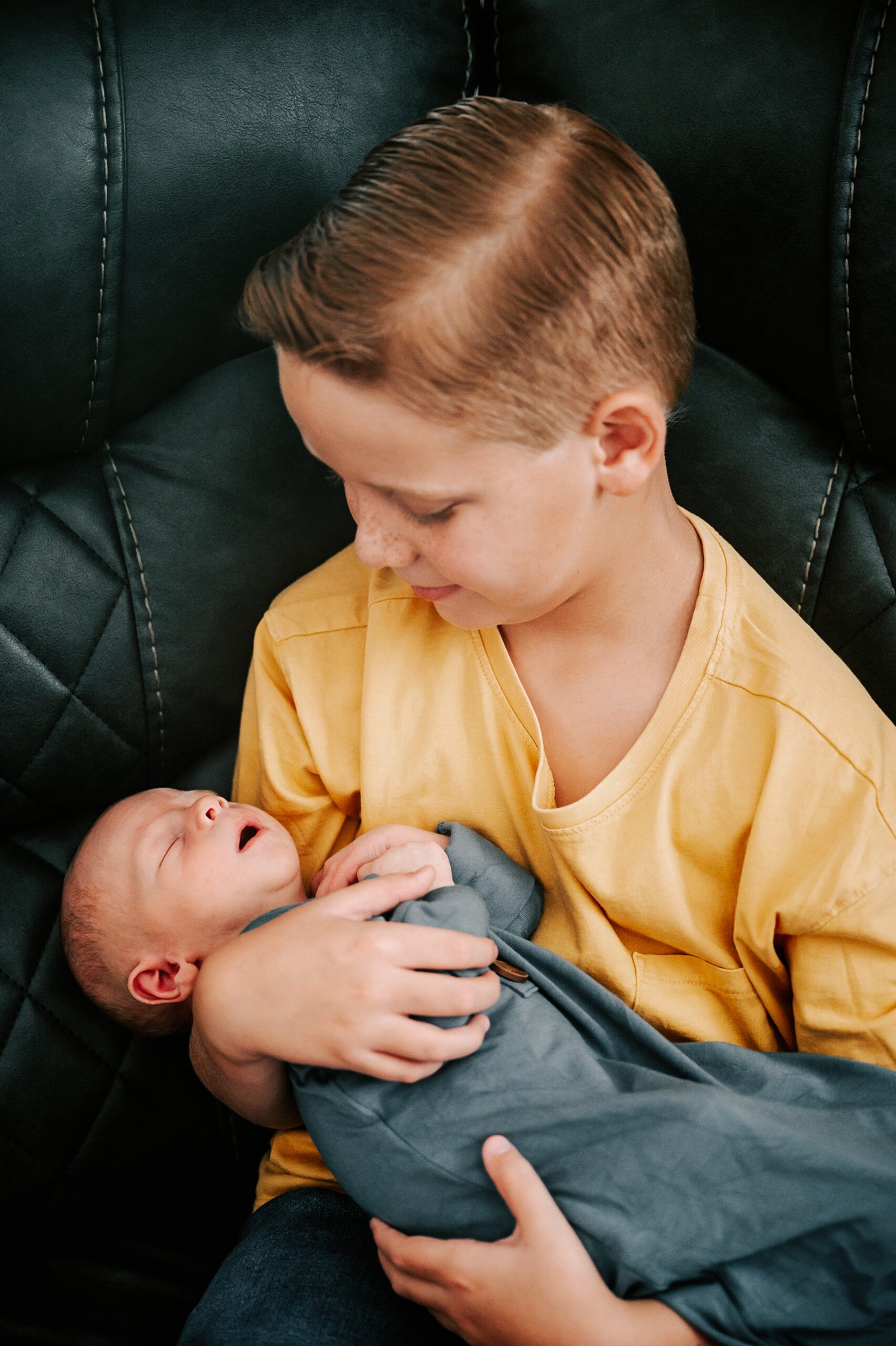 A young boy in a yellow shirt holds his sleeping newborn baby brother in his arms while sitting on a black couch