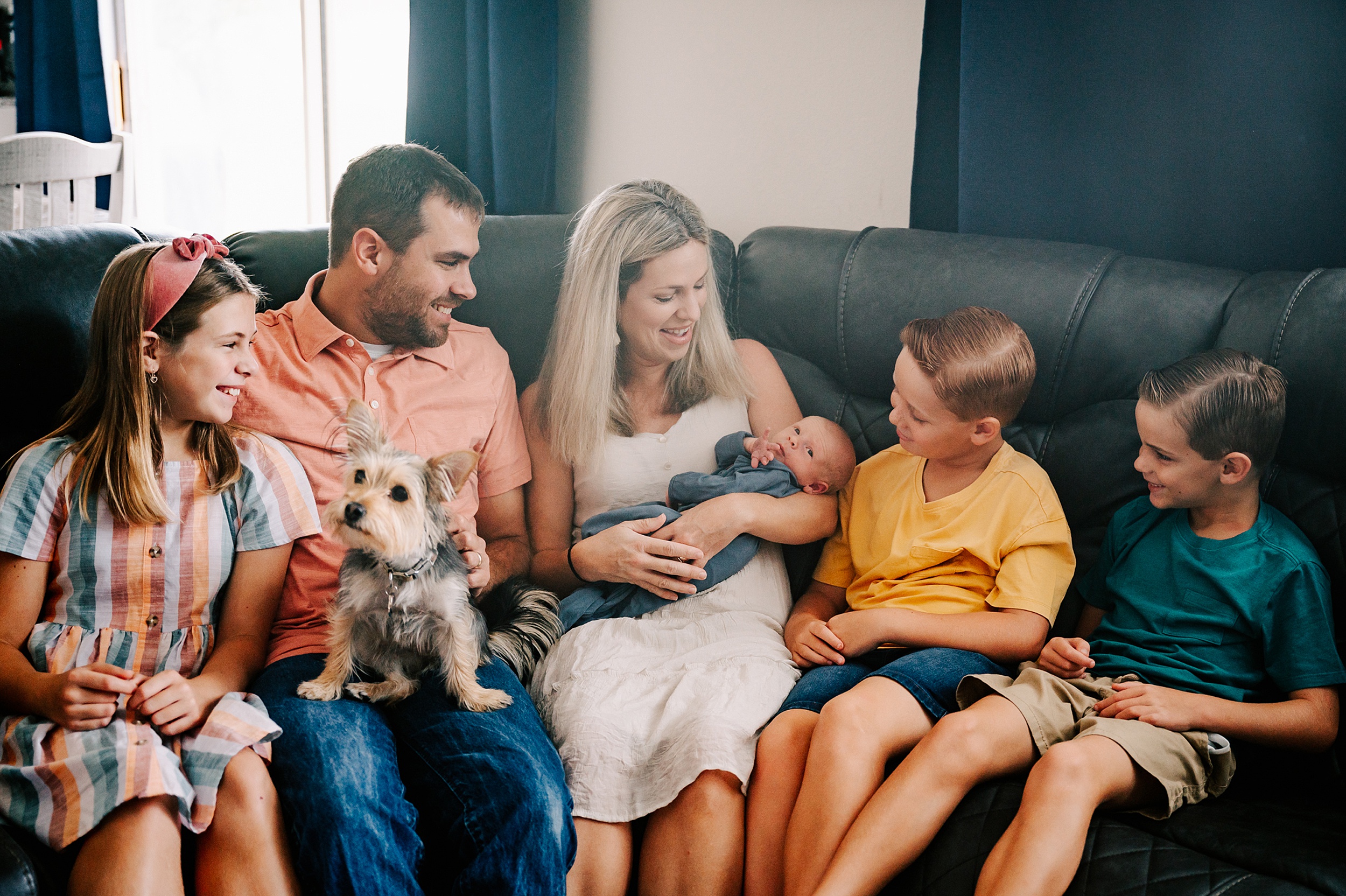 A family of six sit on a couch together looking at the newborn baby in mom's arms with the puppy on dad's lap