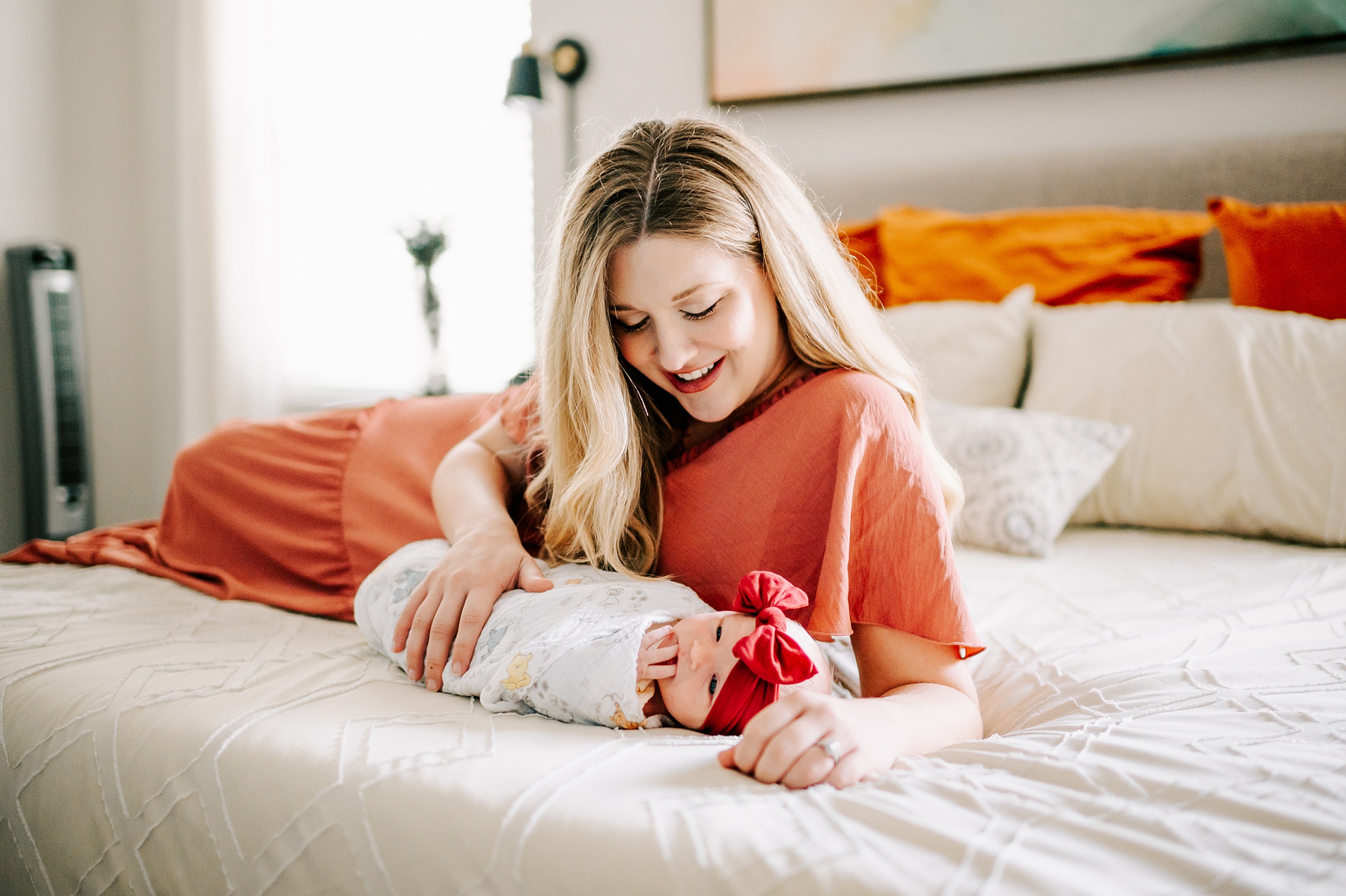 A mother in a red dress lays across a bed while cuddling with her newborn baby in a swaddle and red bow carolinas fertility institute charlotte