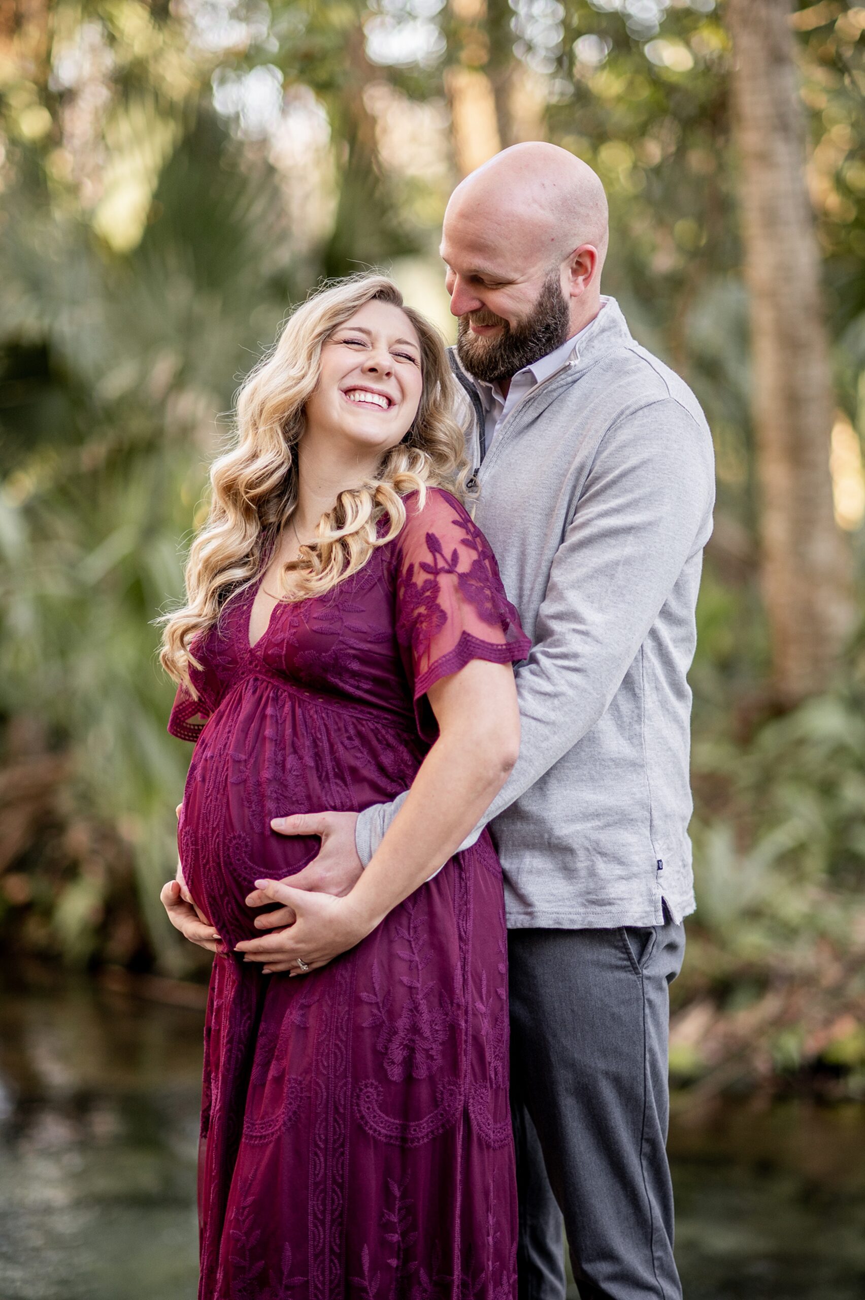 A mom to be is hugged from behind by her husband while wearing a purple lace maternity gown