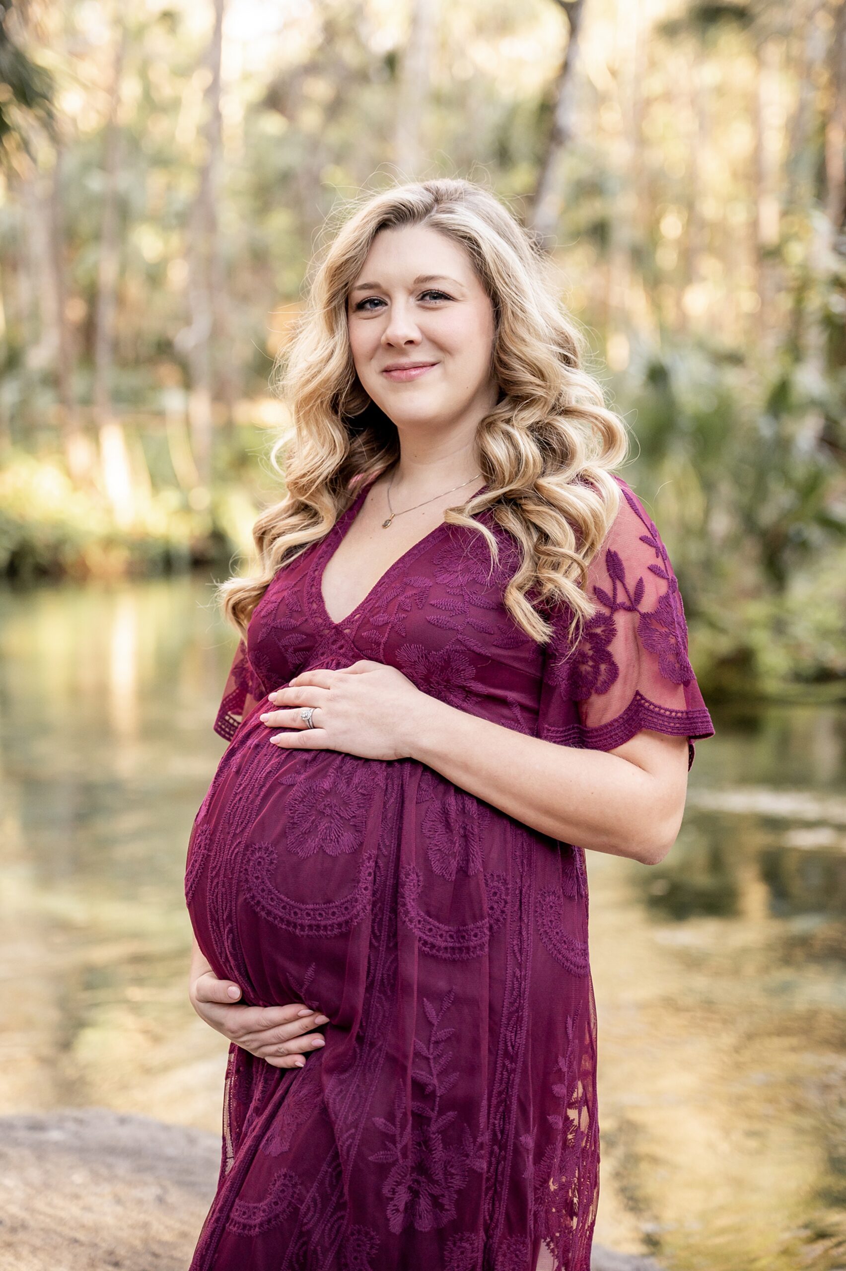 A mom to be in a purple lace maternity gown stands by a river holding her bump carrboro midwifery