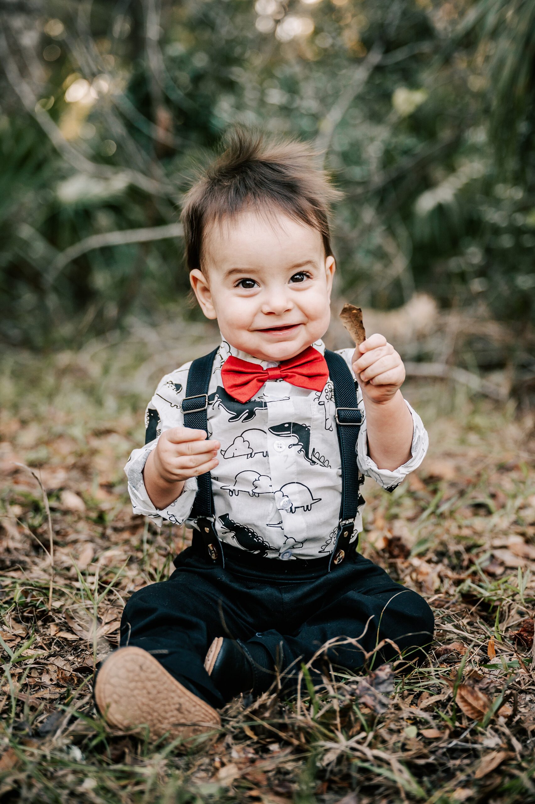 A young toddler in suspenders and a red bowtie sits in a forest while playing with leaves