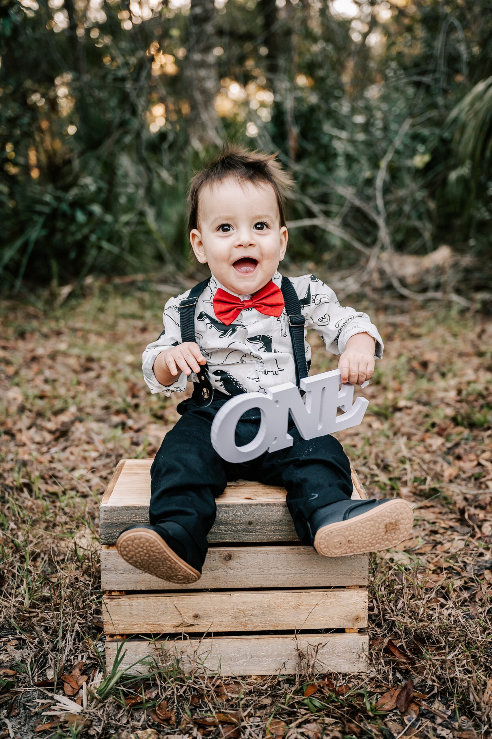 A one year old boy in black pants, suspenders, dinosaur button down shirt and red bowtie plays with a wooden one and sitting on a wooden crate in a forest childtime of greensboro