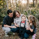 A mother and father squat in a forest while helping their toddler son stand and walk with their older daughter at his side childtime of greensboro