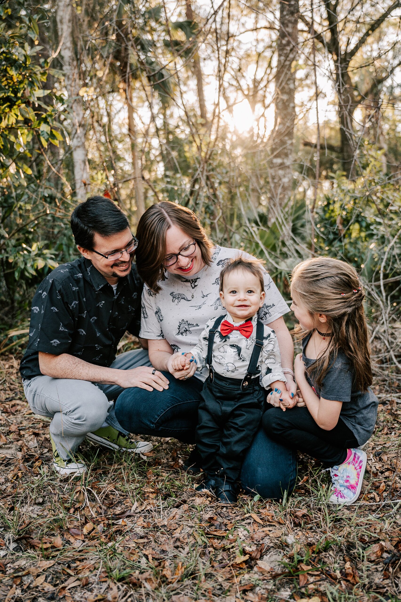 A mother and father squat in a forest while helping their toddler son stand and walk with their older daughter at his side childtime of greensboro