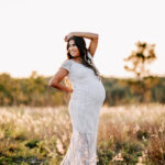 A mother to be in a white lace maternity gown stands in a field of tall golden grass with an arm resting on her head