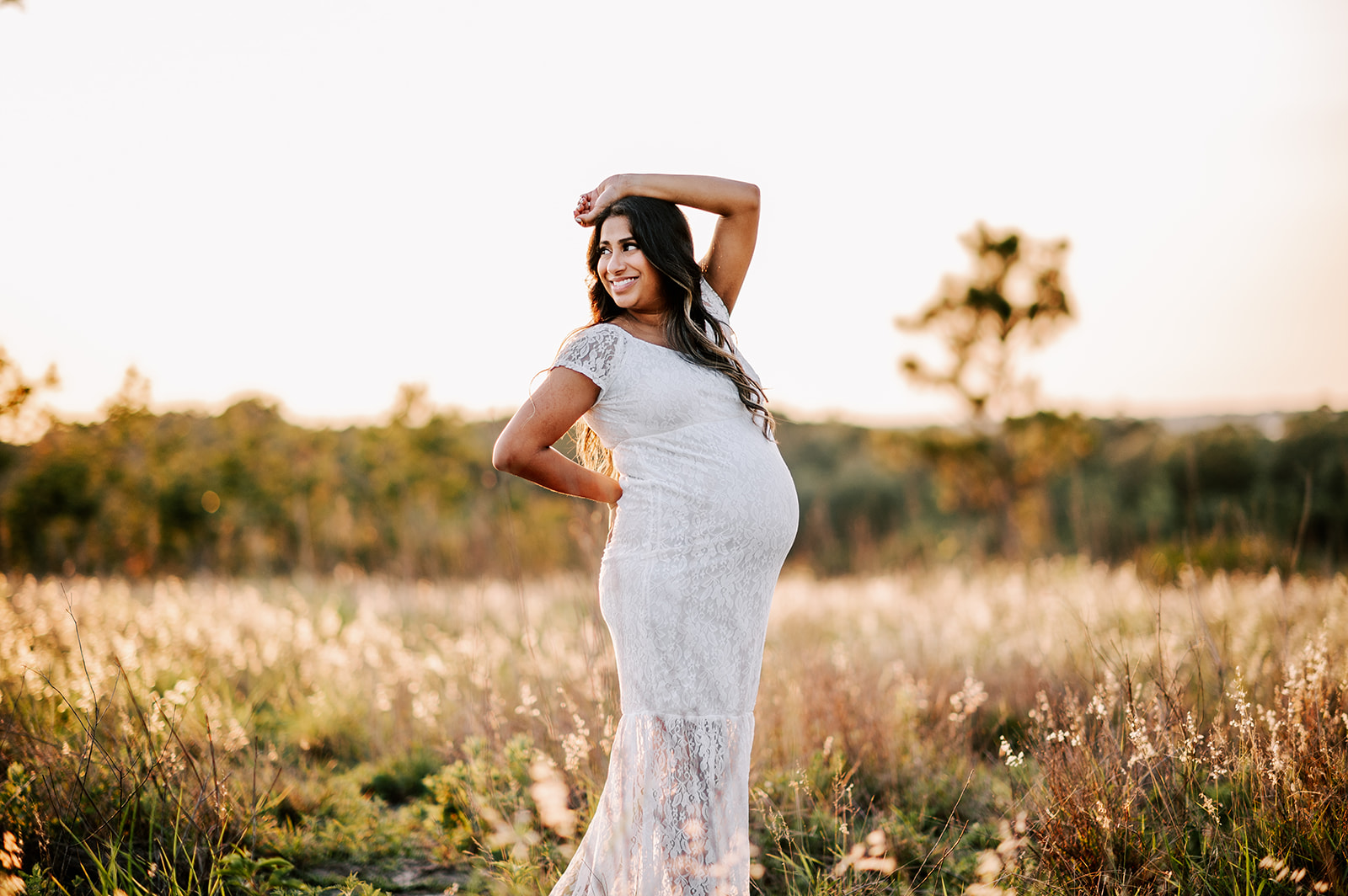 A mother to be in a white lace maternity gown stands in a field of tall golden grass with an arm resting on her head