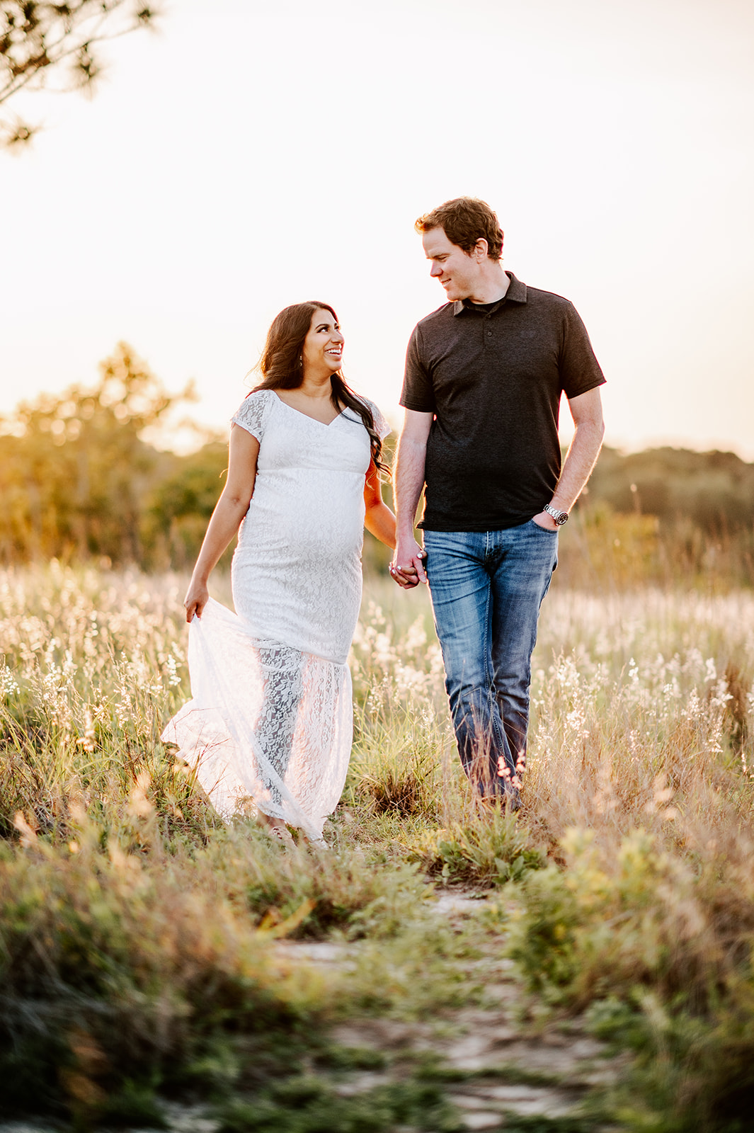 A mom to be in a white lace maternity gown and her husband in a brown polo walk through a field of tall grass while holding hands City of Oaks Midwifery
