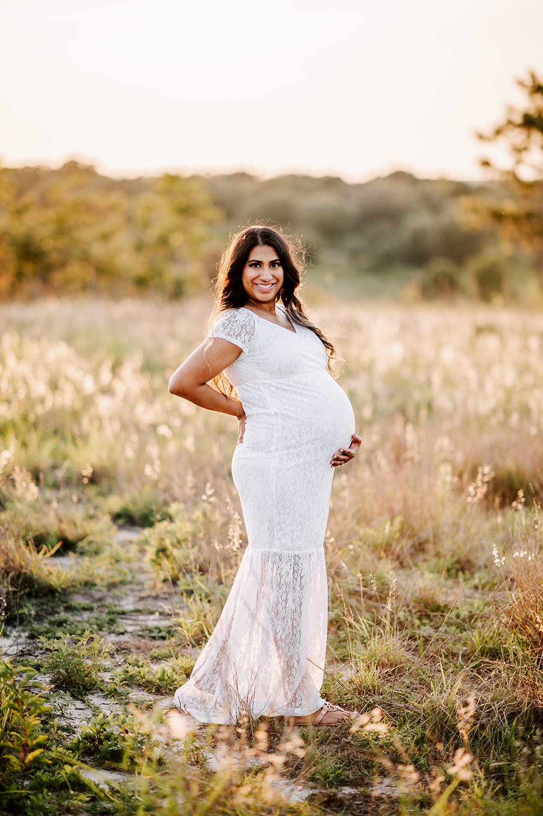 A mother to be in a white lace maternity gown stands in a field of golden grass while holding her bump City of Oaks Midwifery