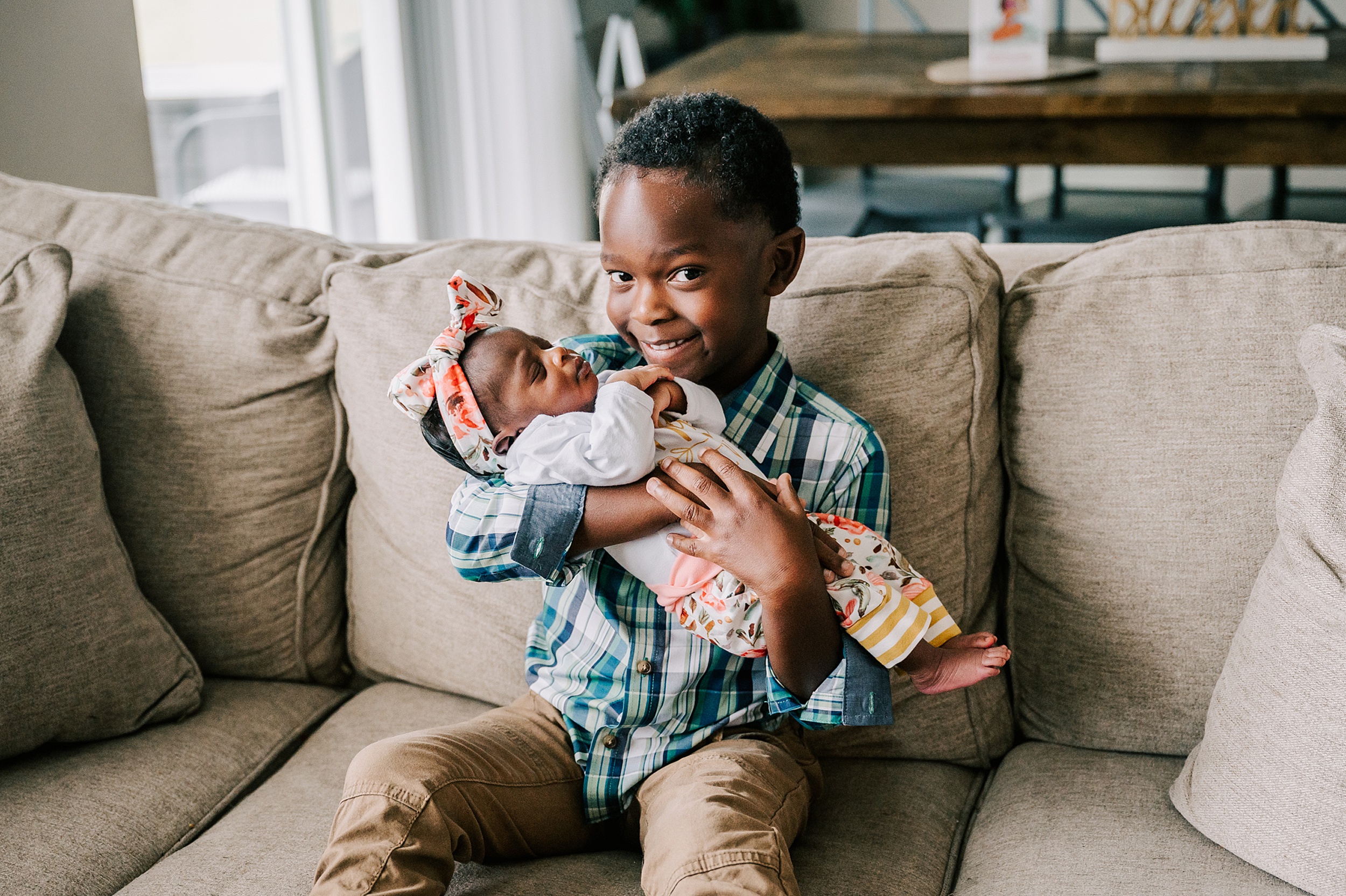 A young boy in a blue plaid shirt holds his newborn baby sister in his arms while sitting on a tan couch diaper bank of north carolina