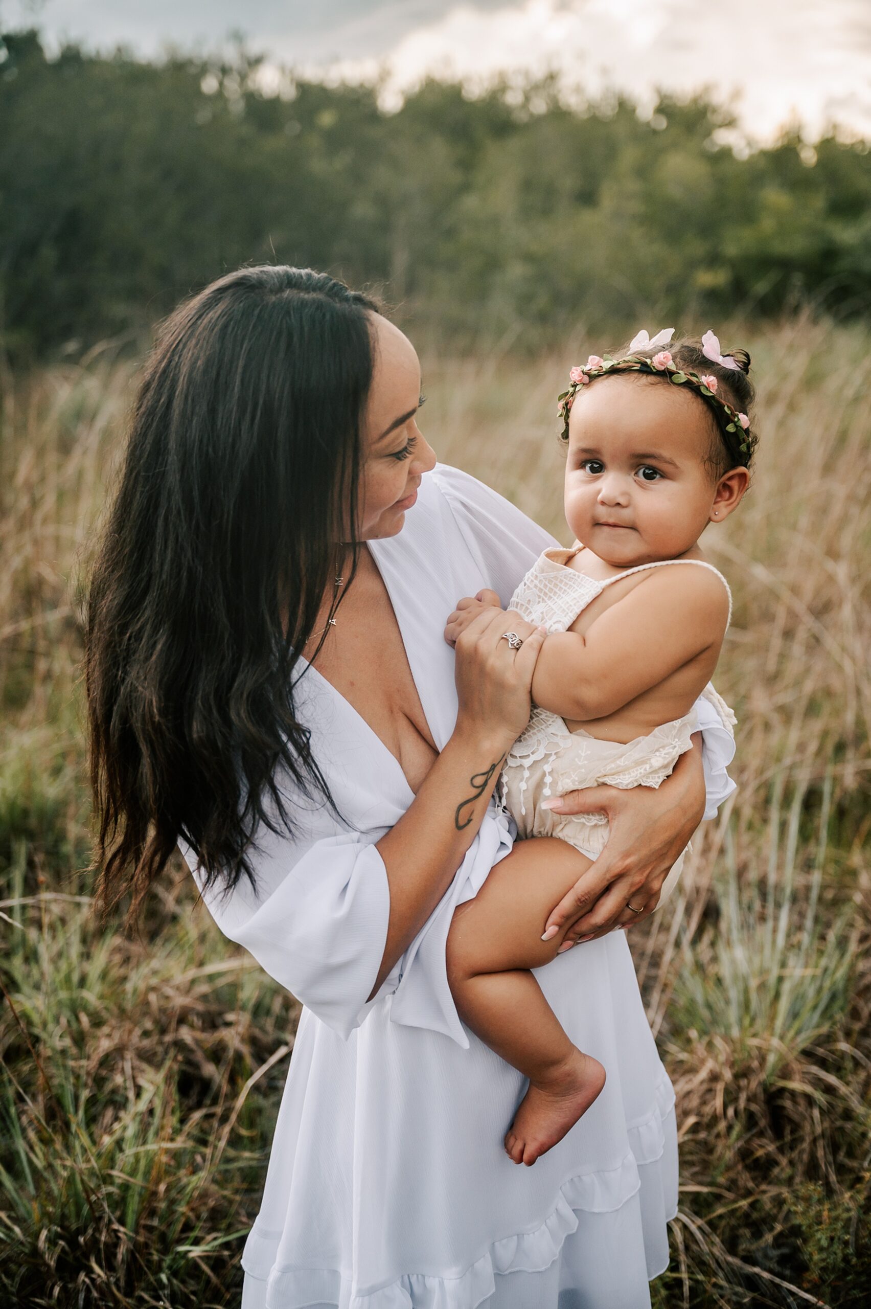 A young mother in a white dress holds her toddler daughter on her hip while standing in a field of golden grass