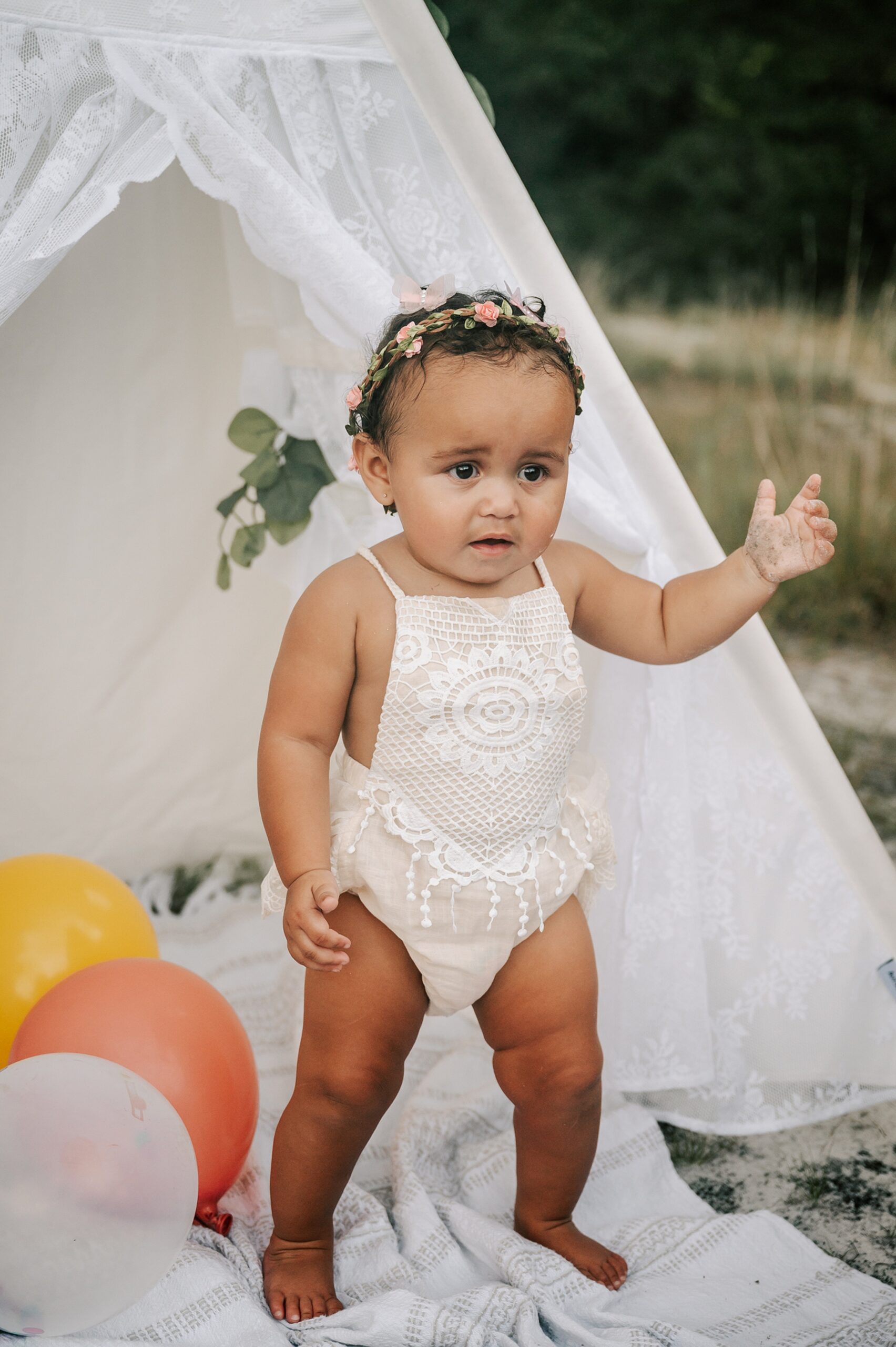 A toddler girl in a lace onesie and floral headband stands on a blanket under a teepee tent with balloons lora belle baby boutique