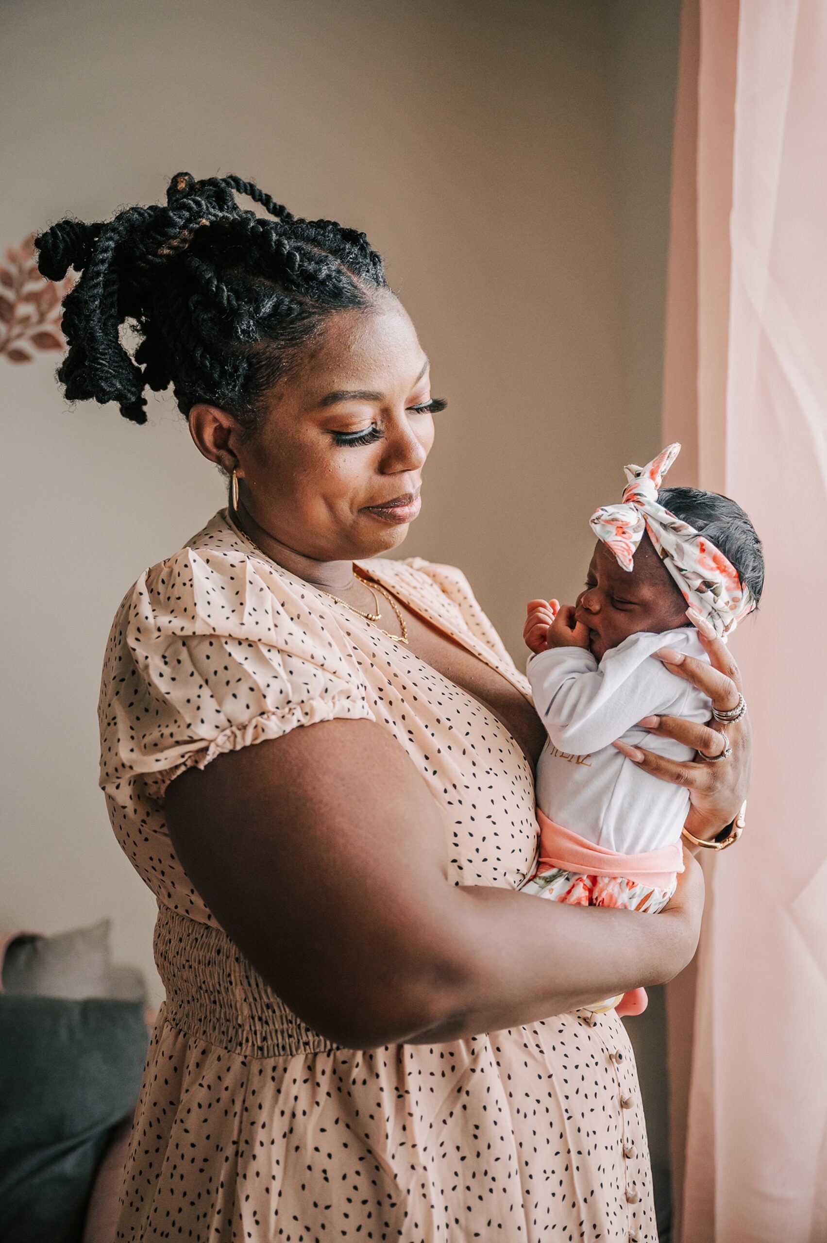 A mother stands by a window with pink curtains while holding her newborn baby daughter in her arms robinhood pediatrics winston salem
