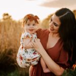 A mother stands in a field of tall golden grass at sunset while holding her young toddler daughter