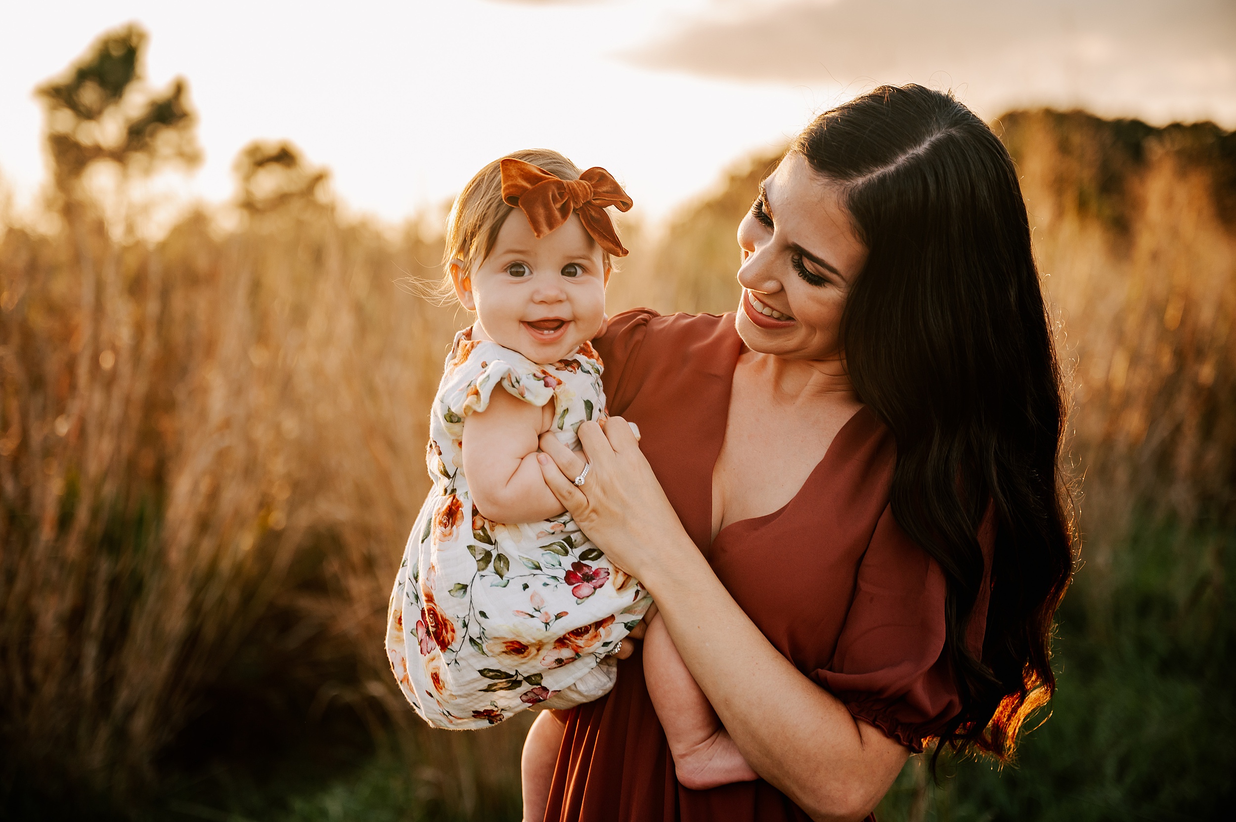 A mother stands in a field of tall golden grass at sunset while holding her young toddler daughter