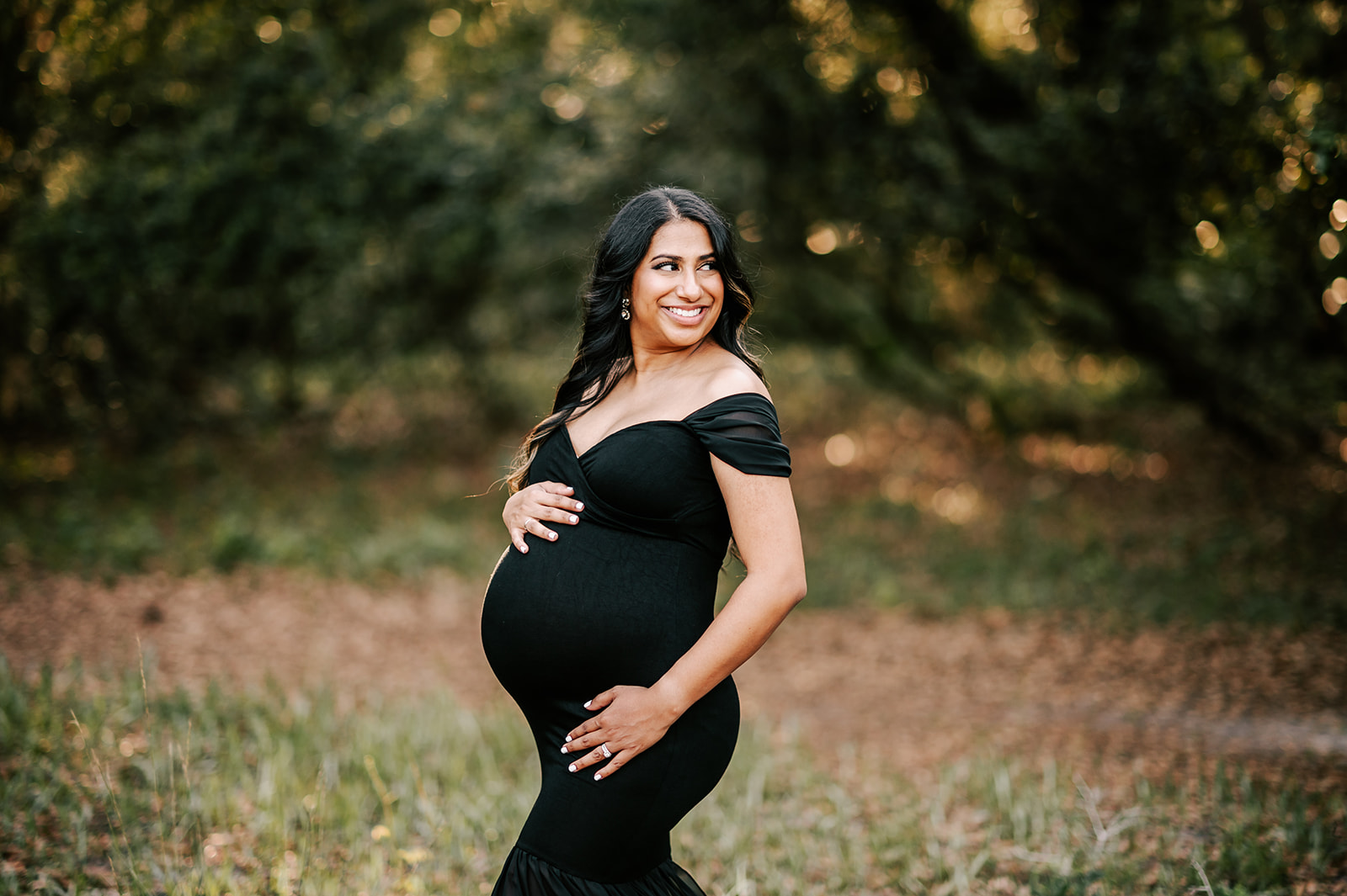 A mom to be stands on the edge of a forest in a black off-the-shoulder maternity gown holding her bump