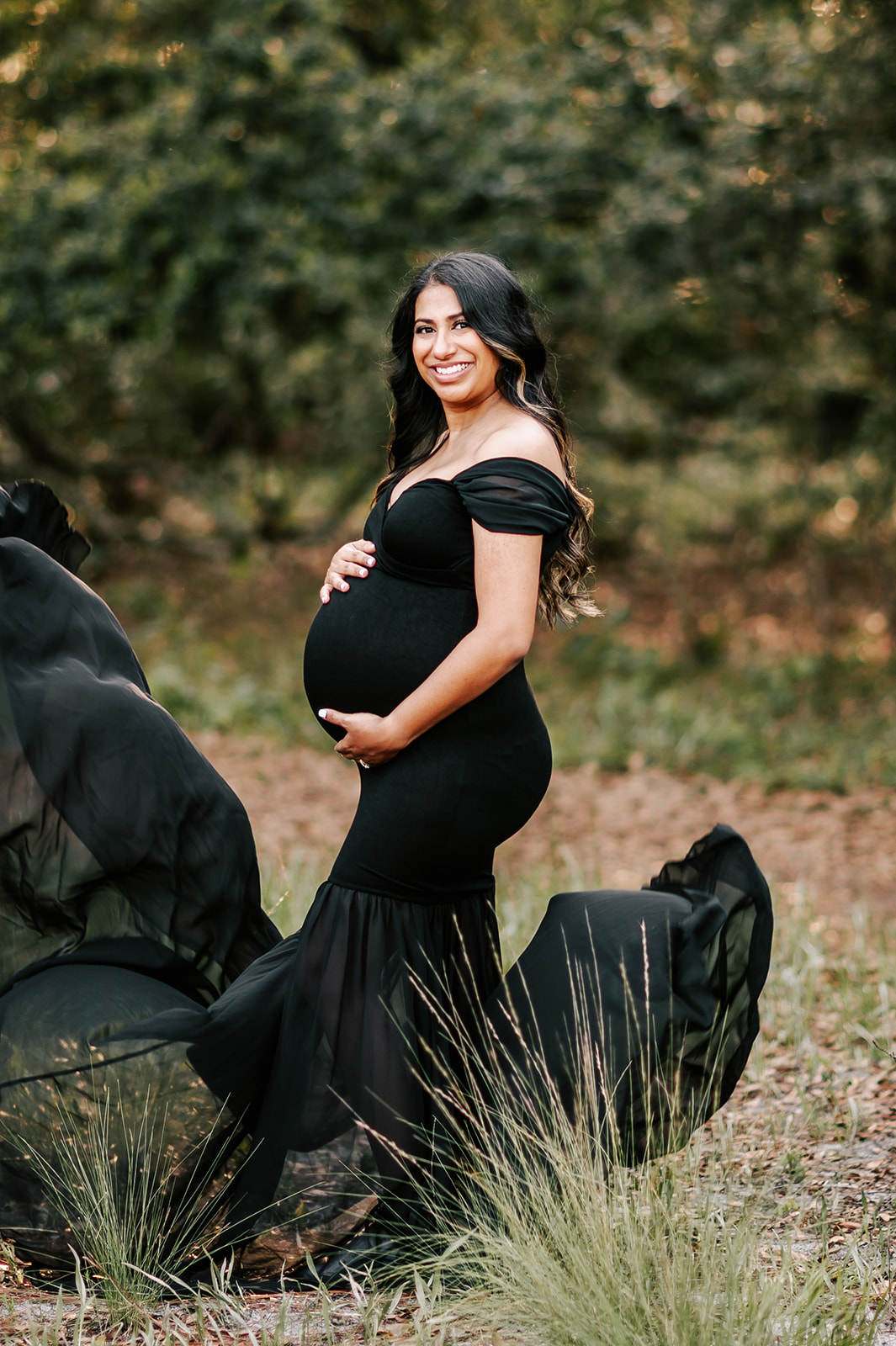 A mother to be stands in a forest in a black maternity gown holding her bump while her train flows in the wind around her What the Bump