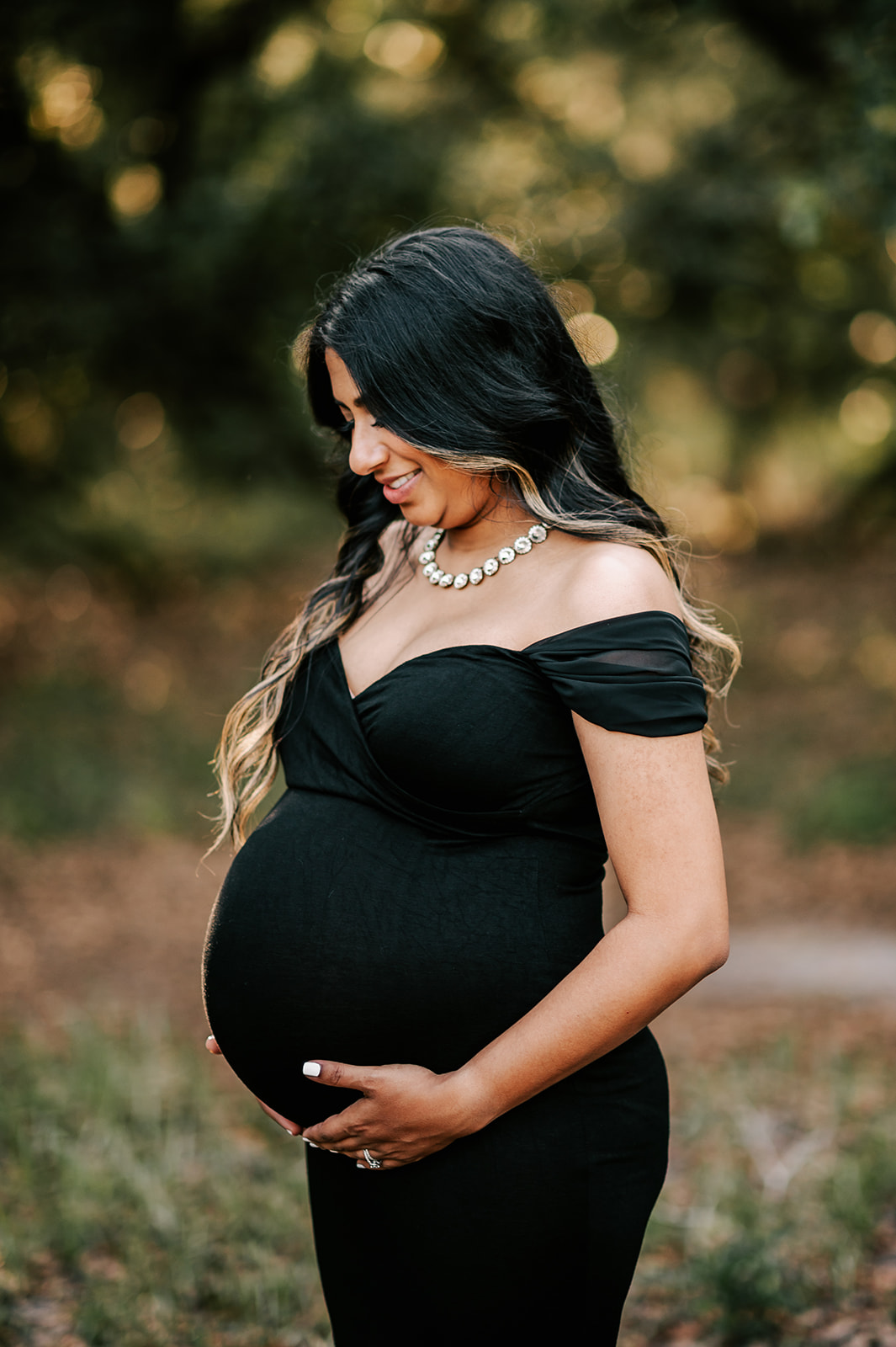 A mom to be in a black maternity gown stands in a forest looking down at her bump What the Bump