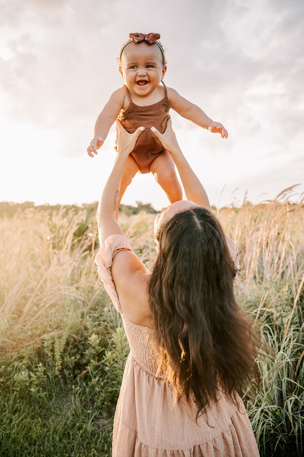 A mother lifts her toddler daughter high over her head while playing in a field of tall grasses at sunset