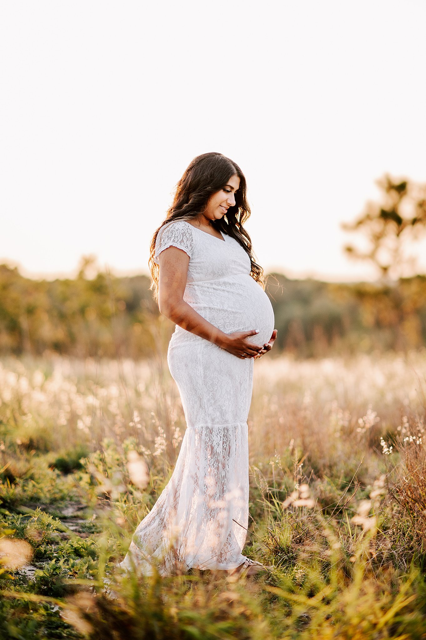A mother to be in a white lace dress stands in a field of tall grass holding her bump