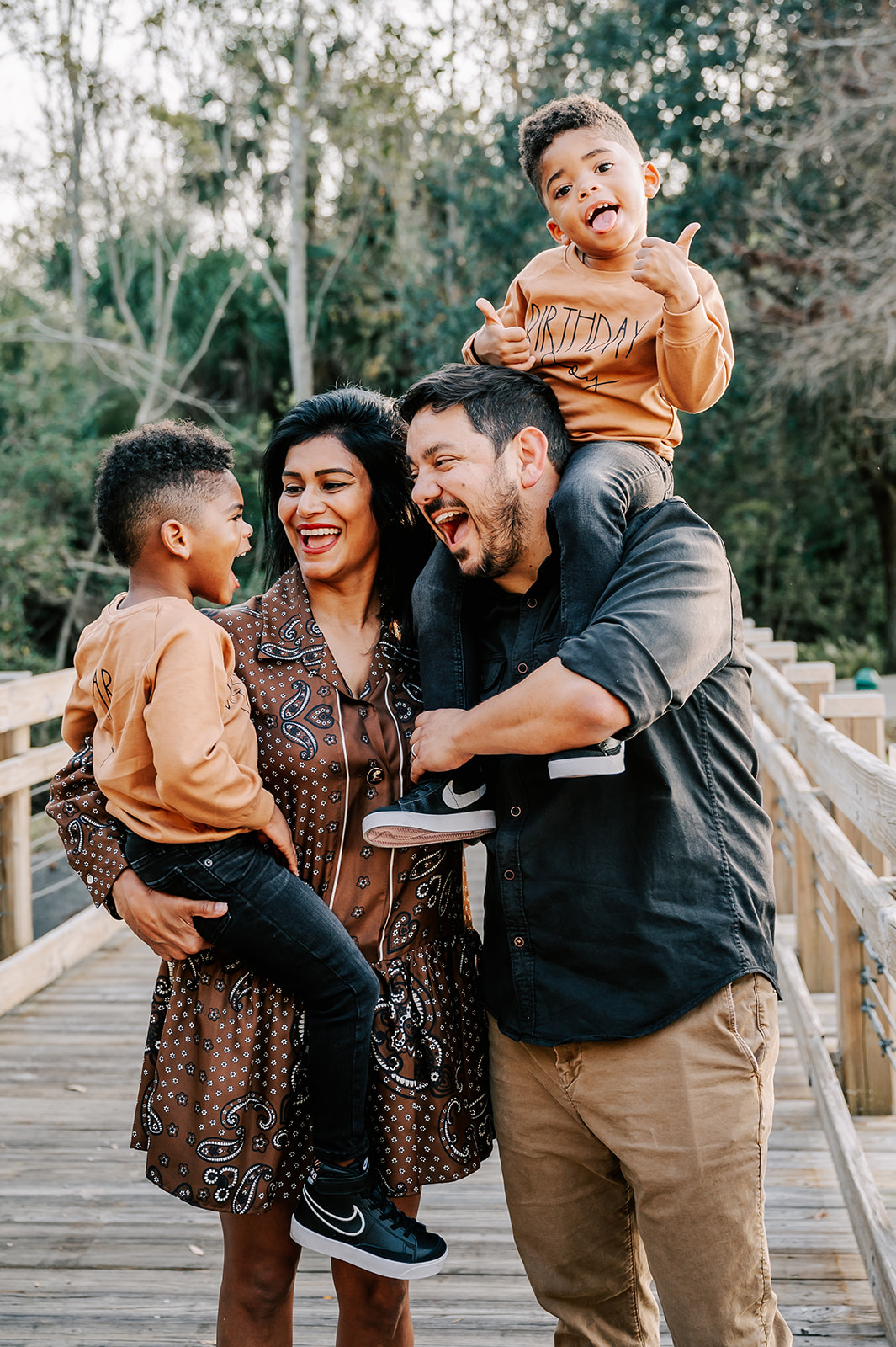 A mother and father play with their twin sons on a wooden park bridge with one on dad's shoulders and other on mom's hip