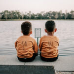 Two young twin boys sit together on the edge of a lake looking over thee water at sunset Sunshine House Early learning Academy