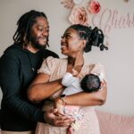 A. other holds her newborn daughter in her arms while dad leans in for a hug standing in their nursery Tiny Toes Prenatal Imaging