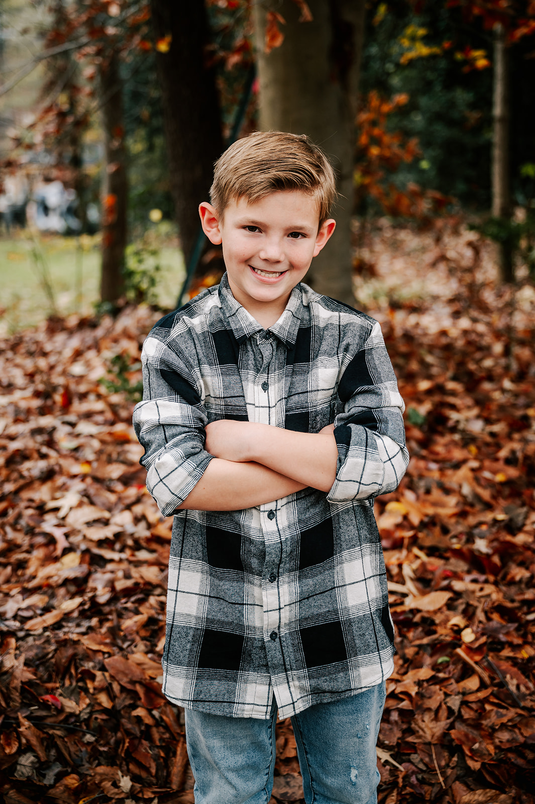 A young boy folds his arms standing in a flannel shirt and jeans in a park