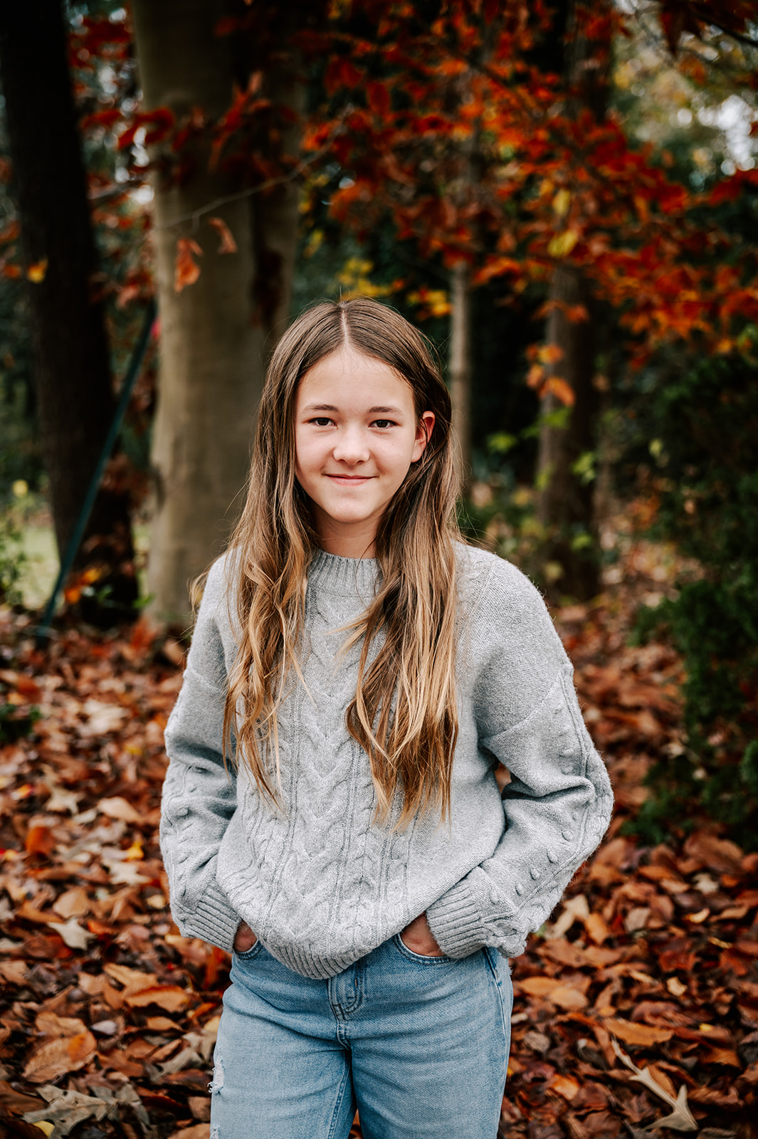 A teenage girl stands with hands in pockets and a grey sweater by a pile of fall leaves Creative Kids Pediatric Dentistry