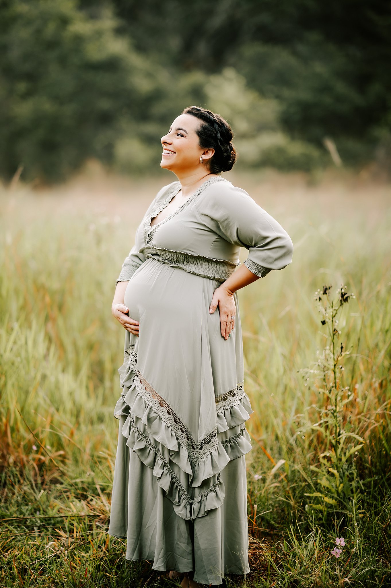 A mother to be in a green maternity dress hoolds her bump and back while standing in a field of tall grasses duke birthing center
