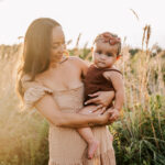 A mother holds her toddler daughter on her hip in a brown dress and brown onesie in a field of tall golden grass at sunset