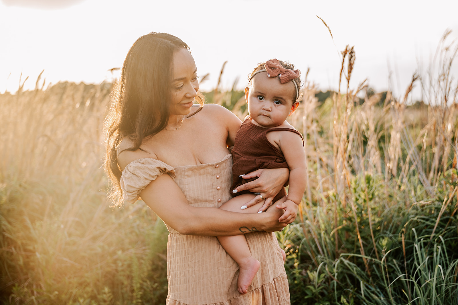 A mother holds her toddler daughter on her hip in a brown dress and brown onesie in a field of tall golden grass at sunset