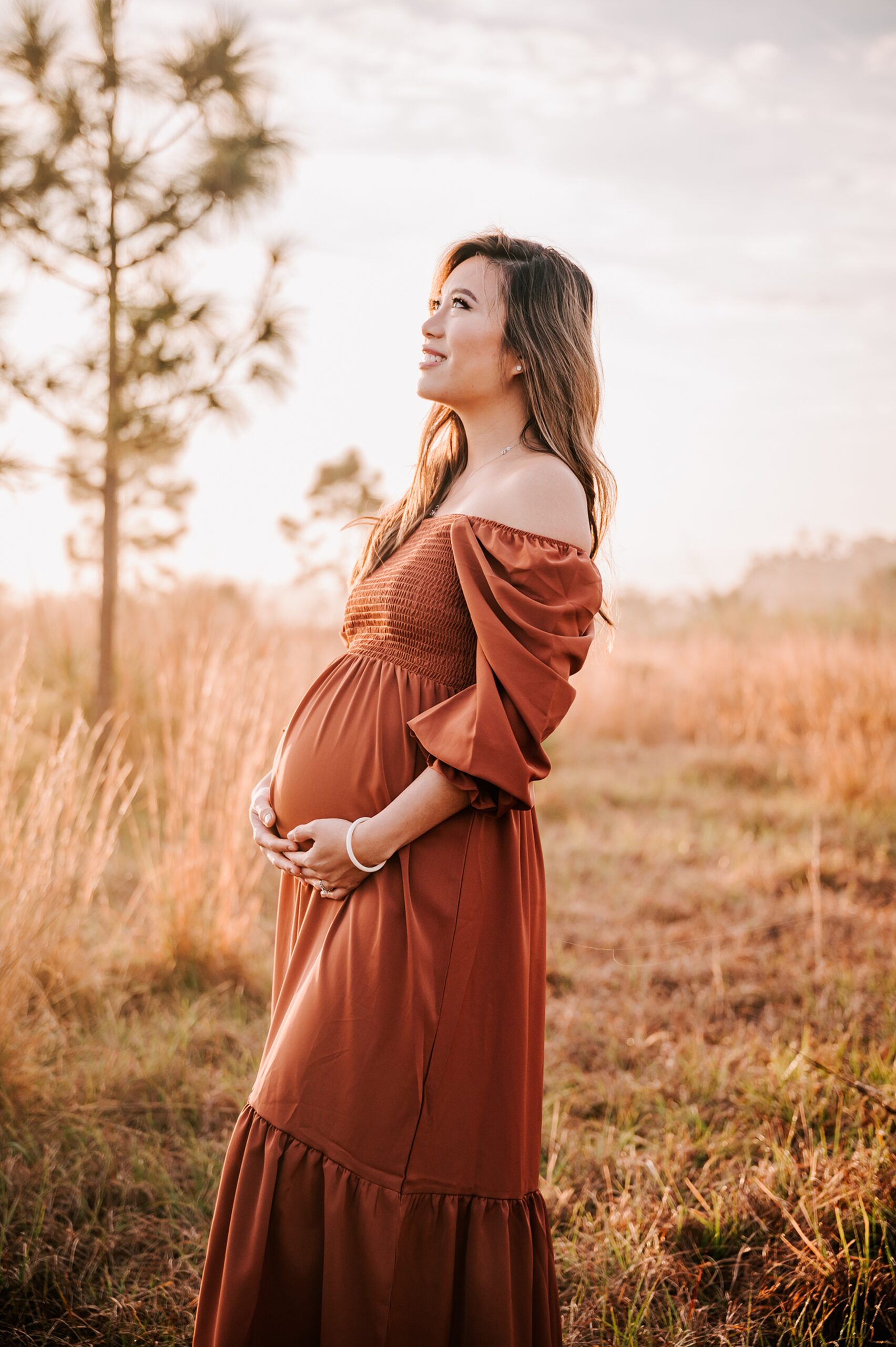 A mother to be holds her bump while looking up at the sky and standing in a field of tall golden grass at sunset wake forest obgyn