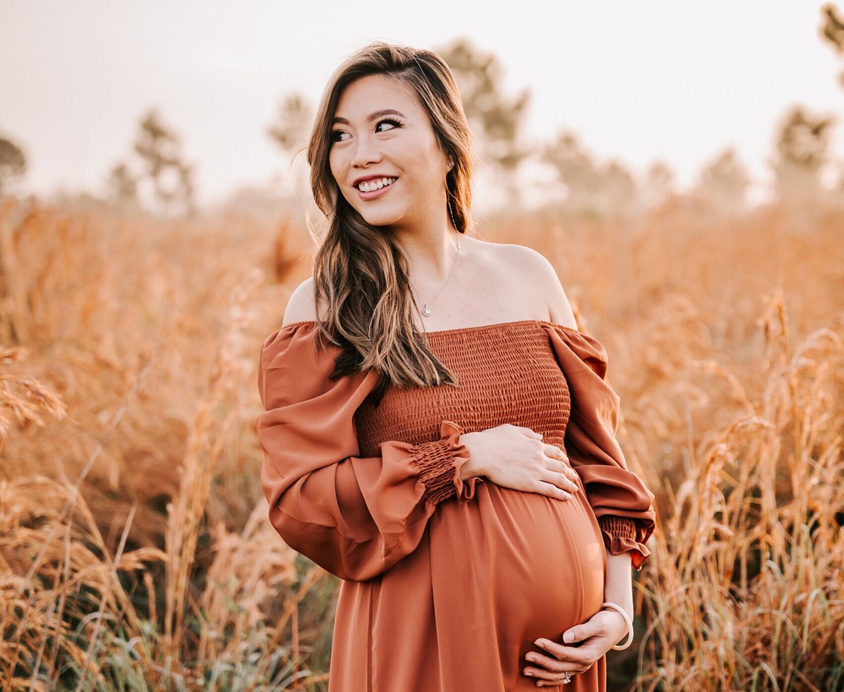 A mother to be holds her bump while looking over her shoulder in a field of tall grass wake forest obgyn