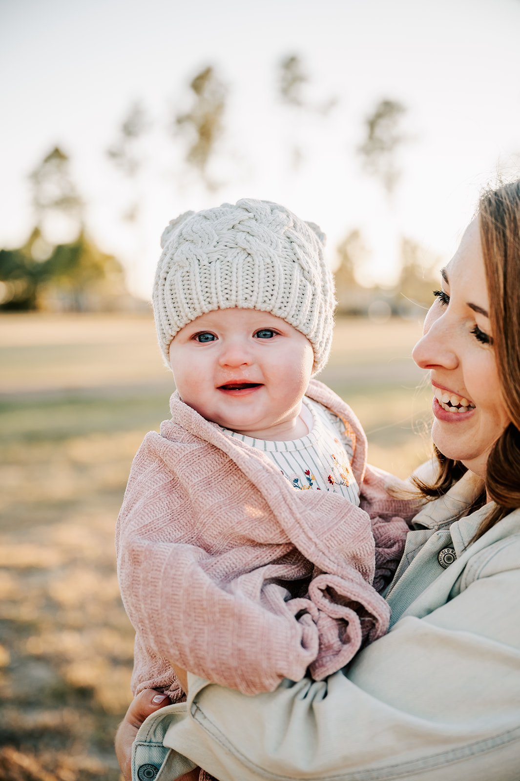 A mother stands in a park at sunset holding her toddler daughter in a pink sweater and knit hat