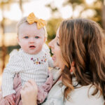 A mother tickles and plays with her young daughter in a white striped onesie in a park Winston Salem Pediatrics