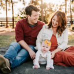 An infant girl sits on a blanket between her parents in a park at sunset