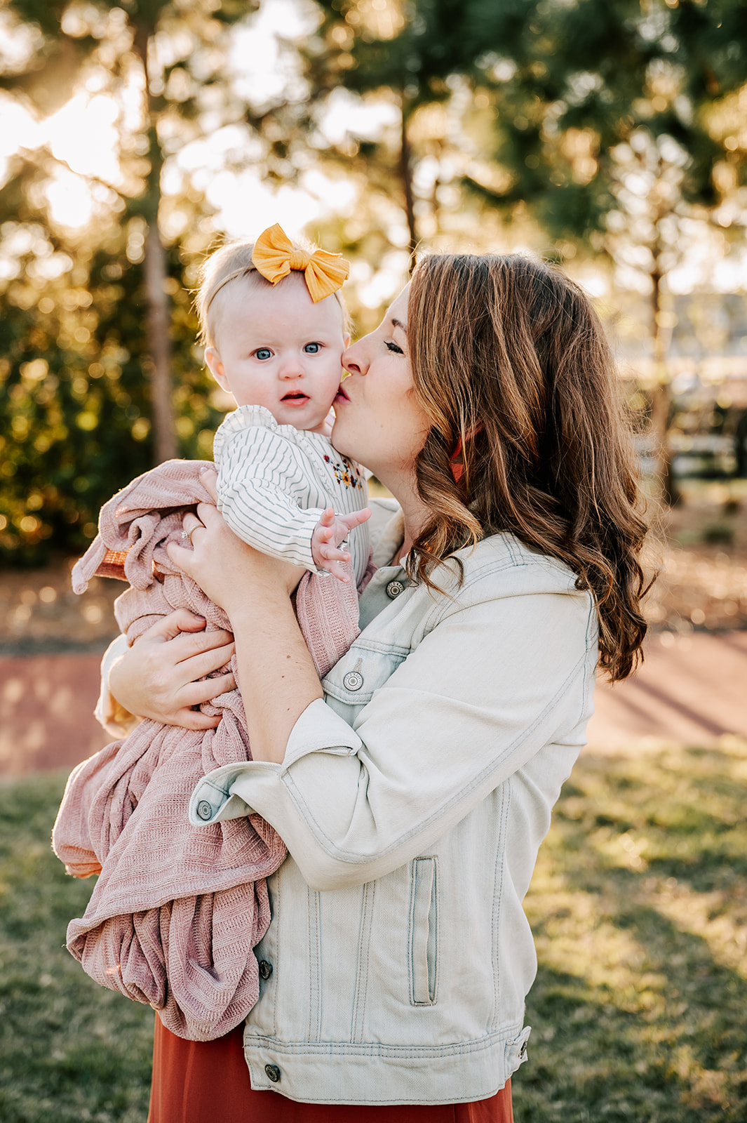 A new mom stands in a park at sunset kissing the cheek of her infant daughter with a yellow bow Charlotte Apple Picking